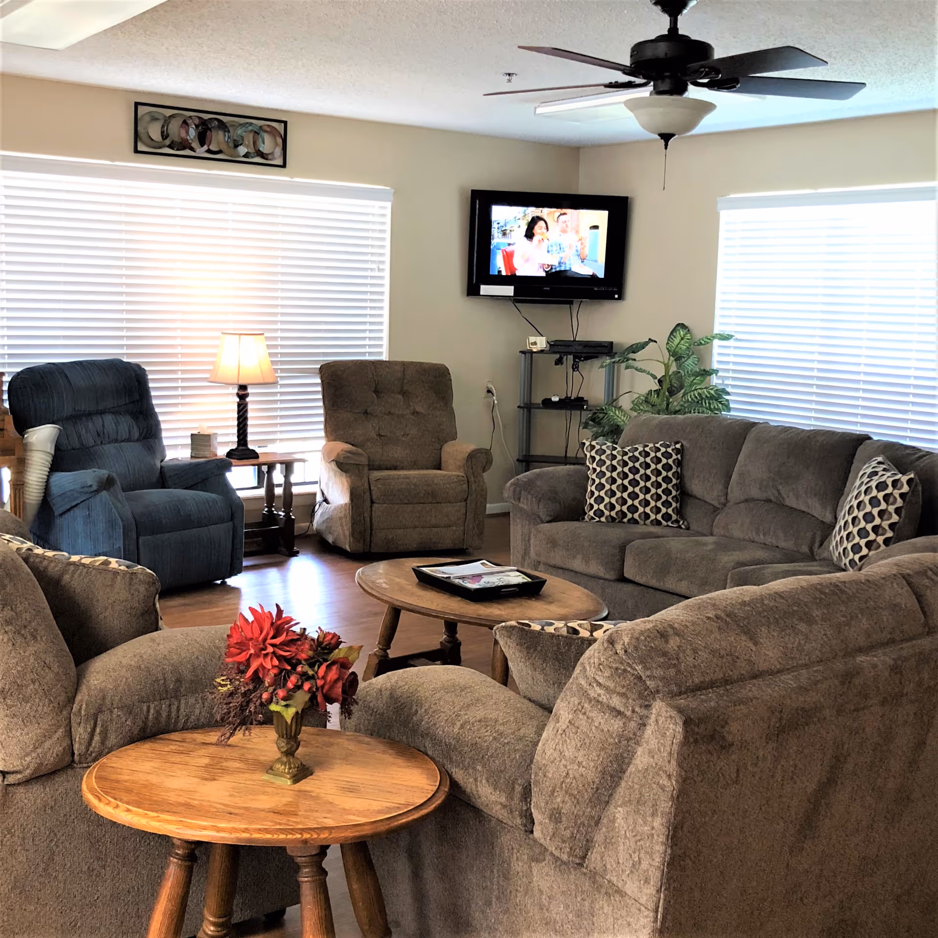 A cozy living room with multiple cushioned armchairs and sofas arranged around a wooden coffee table and side table. A small vase with red flowers is on the side table. The room has two large windows with white blinds, a ceiling fan with a light fixture, a wall-mounted TV showing a program, and a small shelf with electronic devices and a potted plant.