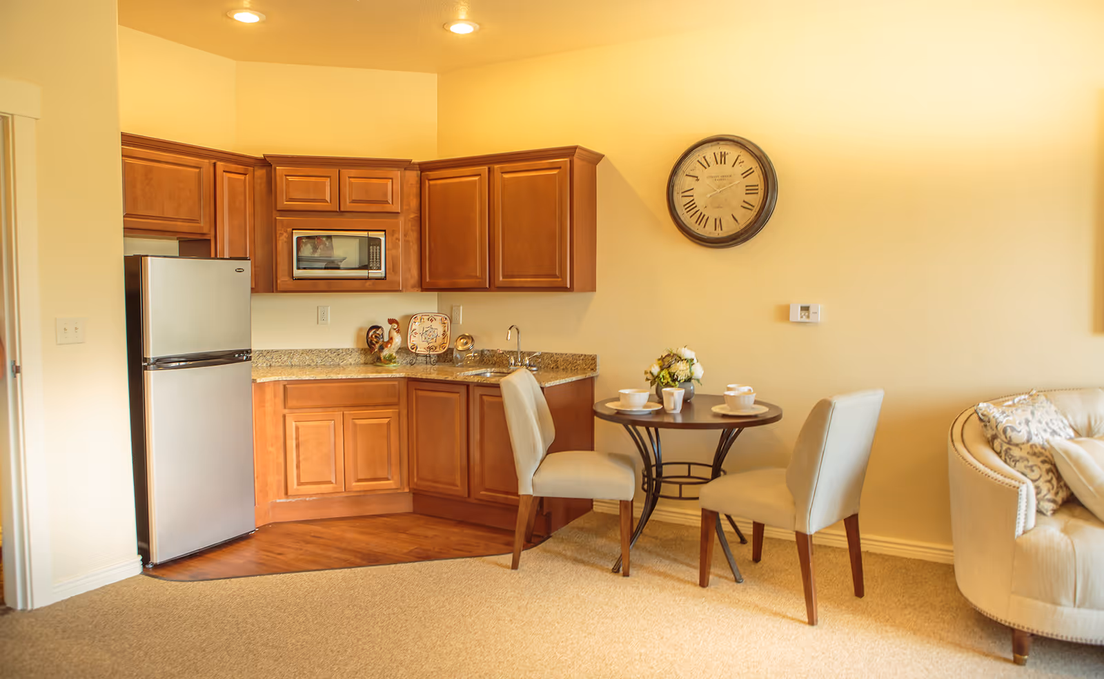 Small open kitchenette with wooden cabinets, a stainless refrigerator, round dining table with two chairs, and a sofa at right.