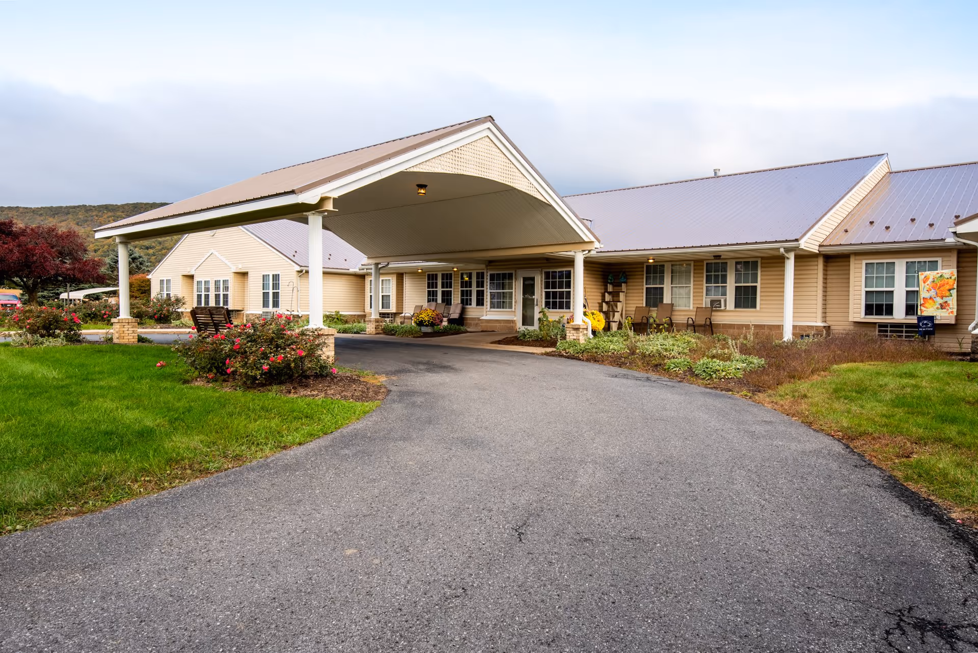 Exterior view of Wynwood House facility showing a single-story building with beige siding and a metal roof. There is a covered driveway entrance with white pillars, surrounded by green grass, flower beds, and outdoor seating. The sky is cloudy with hills in the background.