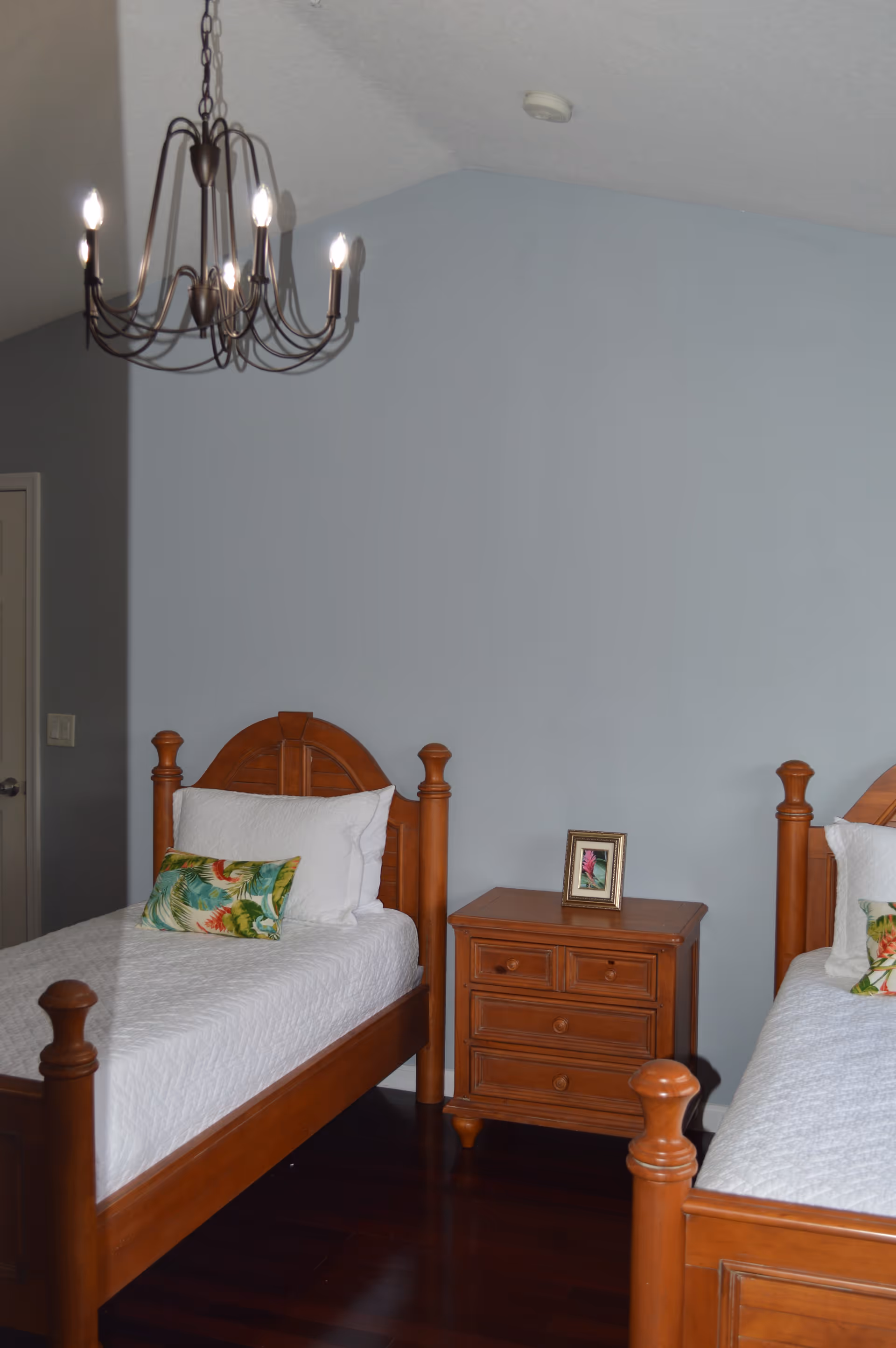 Two wooden twin beds with a nightstand and chandelier in a blue-walled bedroom.