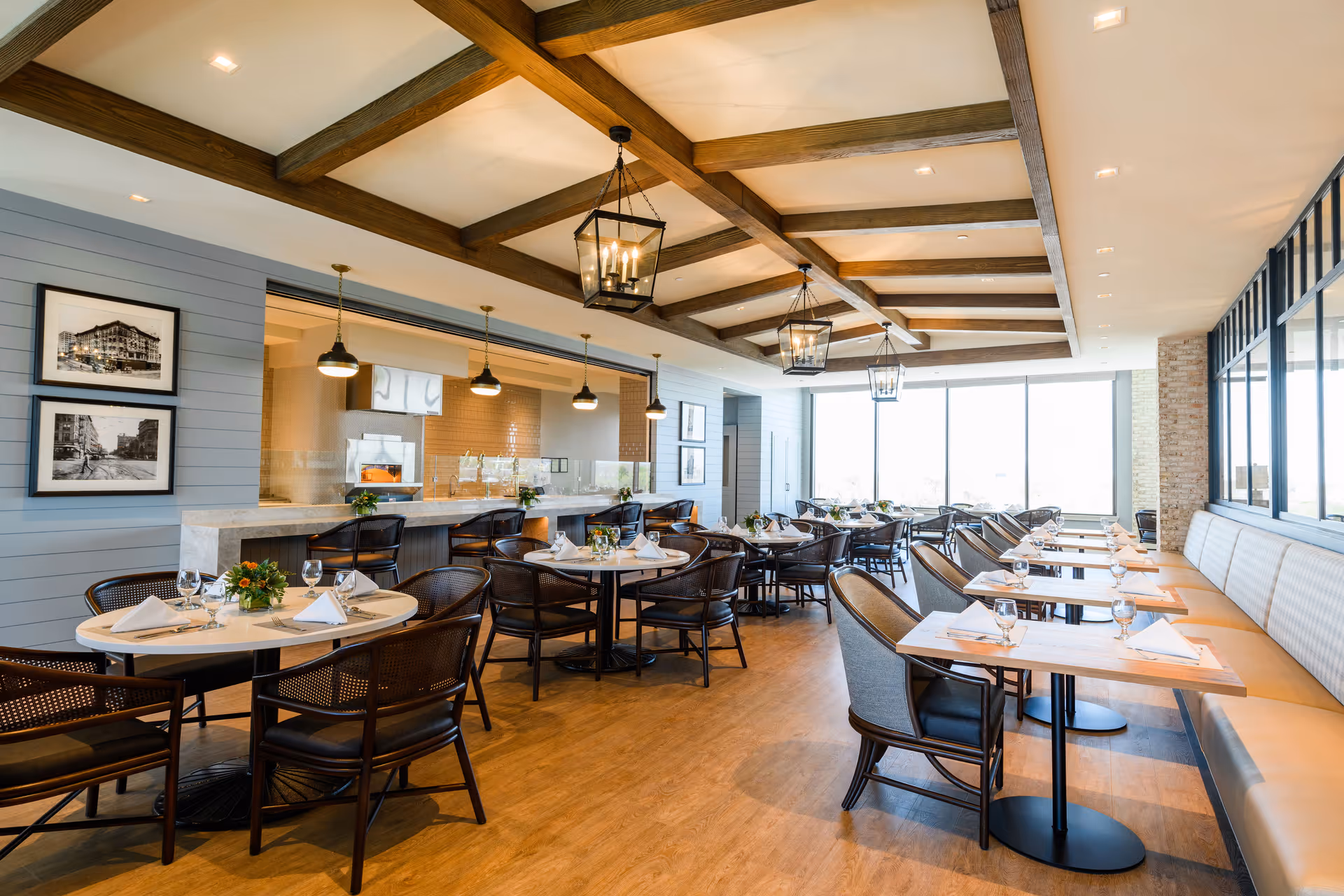 Bright and spacious dining room with wooden beams on the ceiling, multiple round and rectangular tables set with white napkins, glassware, and small floral centerpieces. The room features large windows allowing natural light, a long cushioned bench along one wall, and a counter with bar stools under pendant lights.