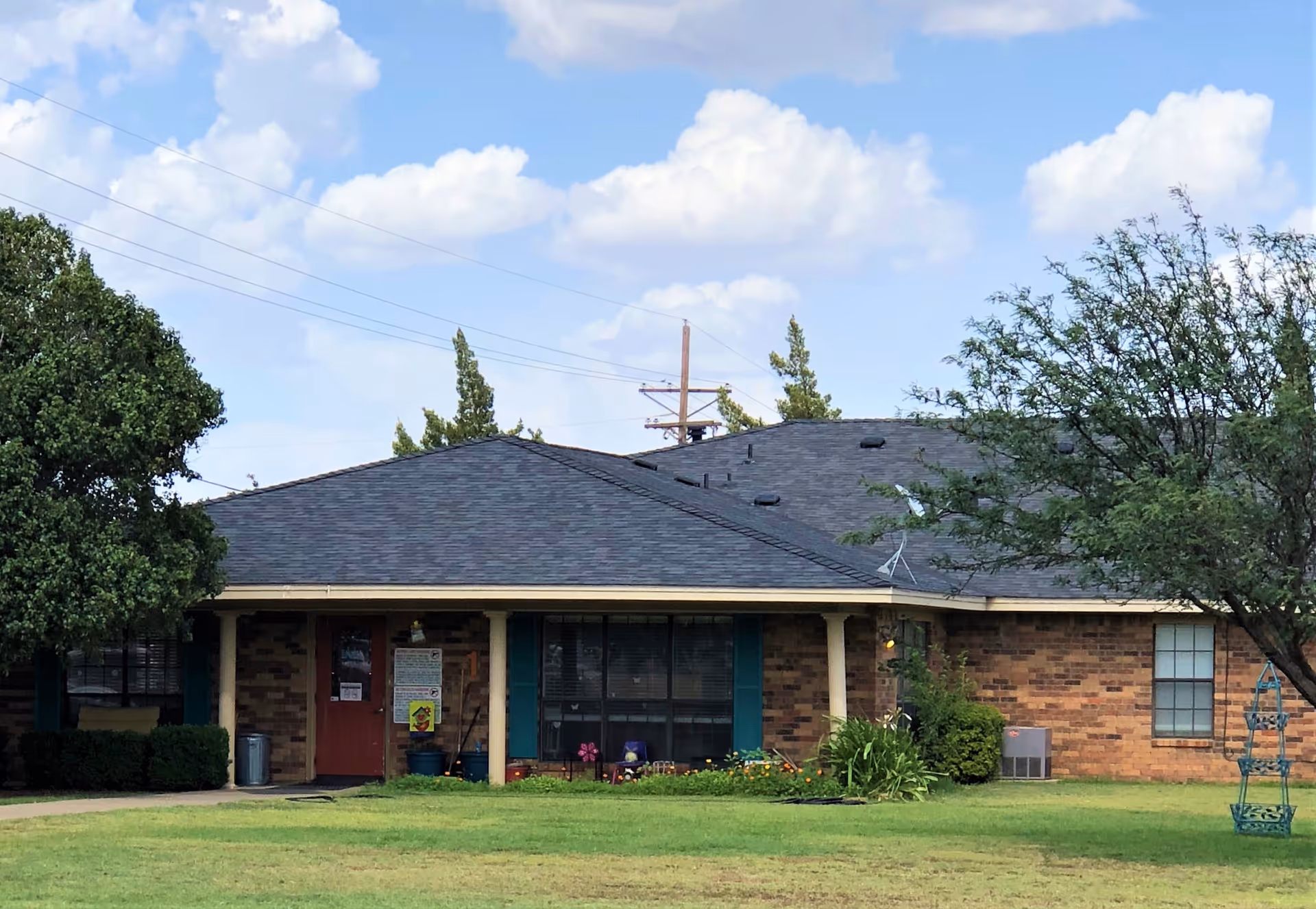 Single-story brick building with a dark shingled roof, green shutters, and a red door. The building is surrounded by a well-maintained lawn with trees and shrubs under a partly cloudy blue sky.