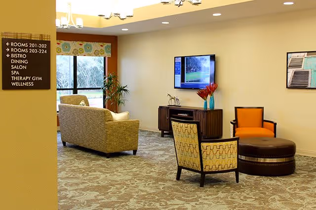 A cozy living room area in a senior living facility with patterned carpet, a beige sofa, two patterned armchairs, an orange armchair, and a round brown ottoman. A flat-screen TV is mounted on the wall above a wooden cabinet with decorative items. A sign on the left wall lists directions to rooms, bistro, salon, spa, therapy gym, and wellness areas. A window with a patterned valance lets in natural light.