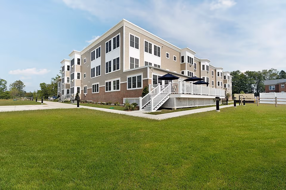 Exterior view of a modern three-story senior living facility building with beige and white siding and brick accents. The building features multiple windows and a raised deck with white railings and dark patio umbrellas. The surrounding area includes a well-maintained green lawn and a paved walkway under a partly cloudy sky.