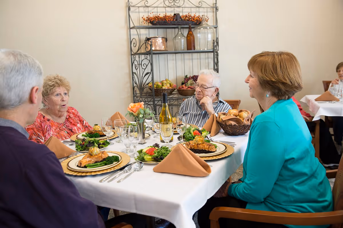 A group of older adults seated around a dining table in a senior living dining room with plated meals and table settings.