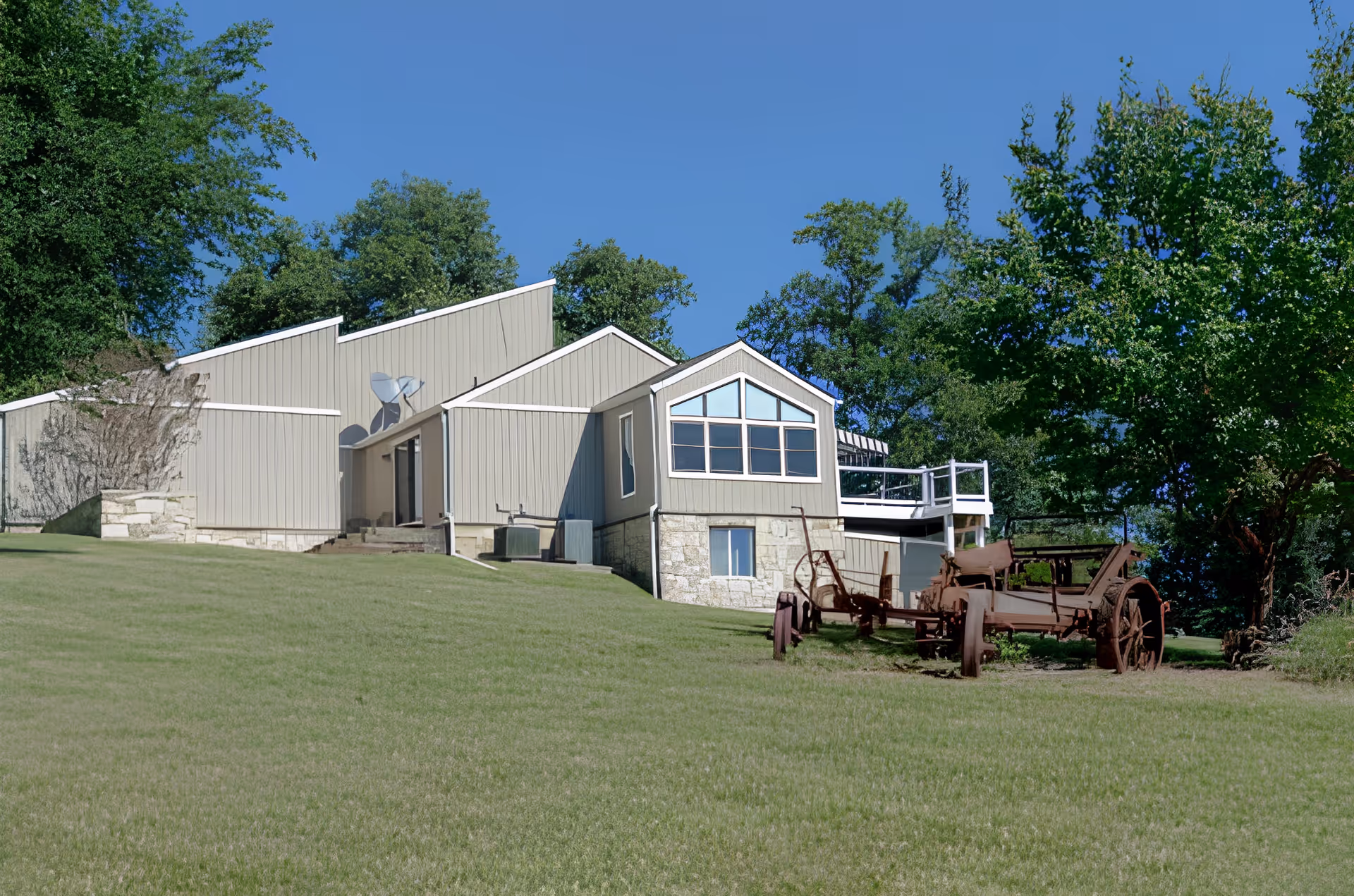 A light-colored building on a grassy slope with trees behind it and antique farm equipment in the foreground.