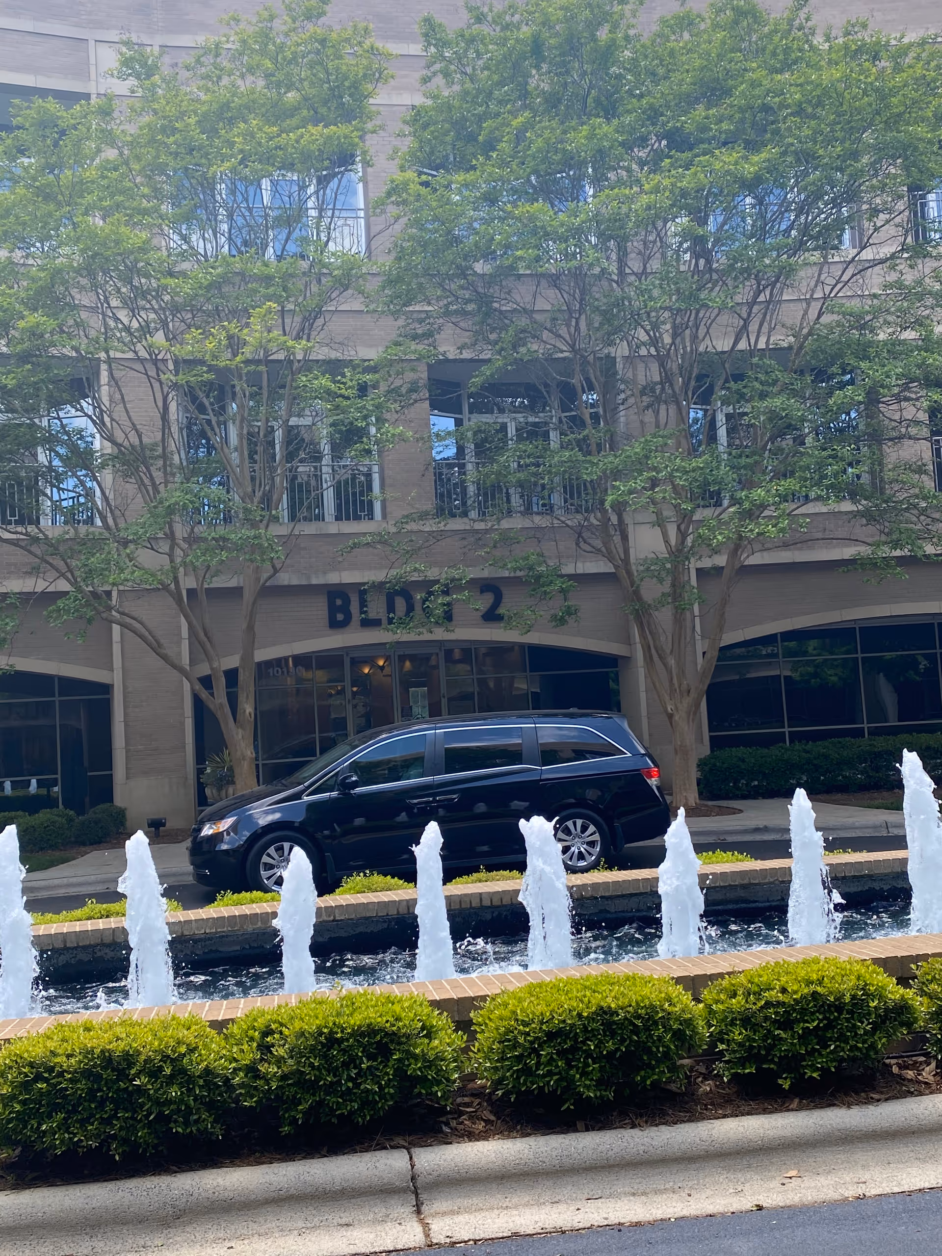 Exterior view of a building labeled BLDG 2 with fountains, trees, and a parked van.