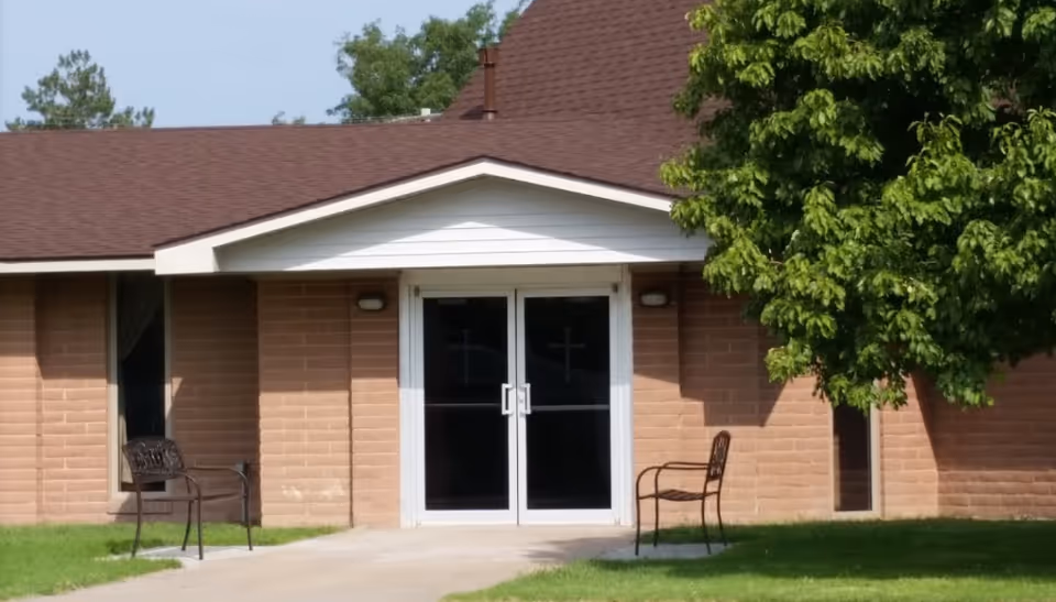 Brick building with double doors, lawn, and metal chairs outside the entrance.
