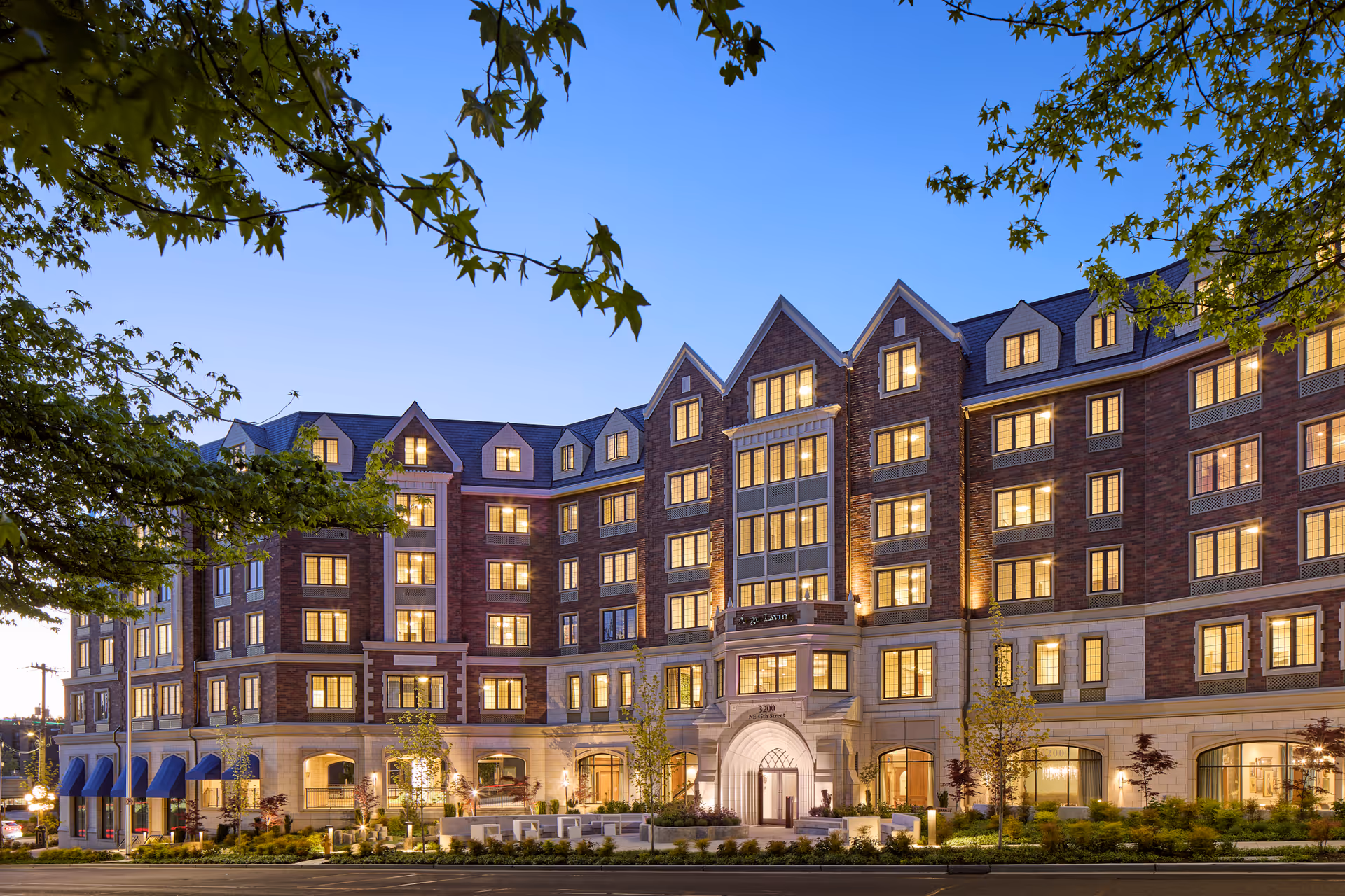 Exterior view of a large, multi-story senior living facility named Aegis Living Laurelhurst during twilight. The building features a brick facade with numerous lit windows, gabled roof sections, and a prominent arched entrance. Trees and landscaping are visible in the foreground under a clear blue sky.