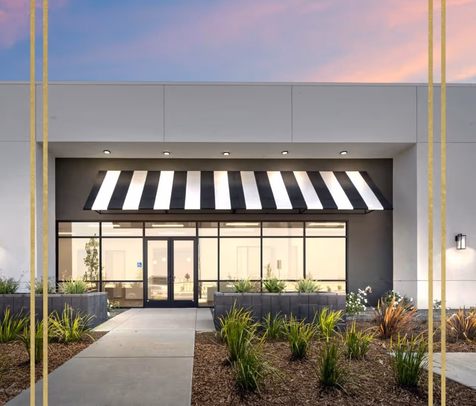 Modern building entrance with glass doors, striped awning, and landscaped entry.