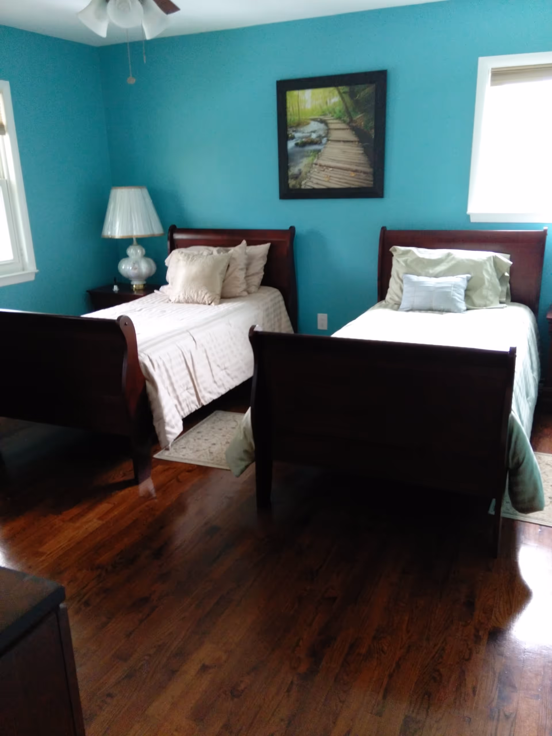 Two twin wooden sleigh beds in a teal-painted bedroom with hardwood floors, a bedside lamp, and framed artwork on the wall.