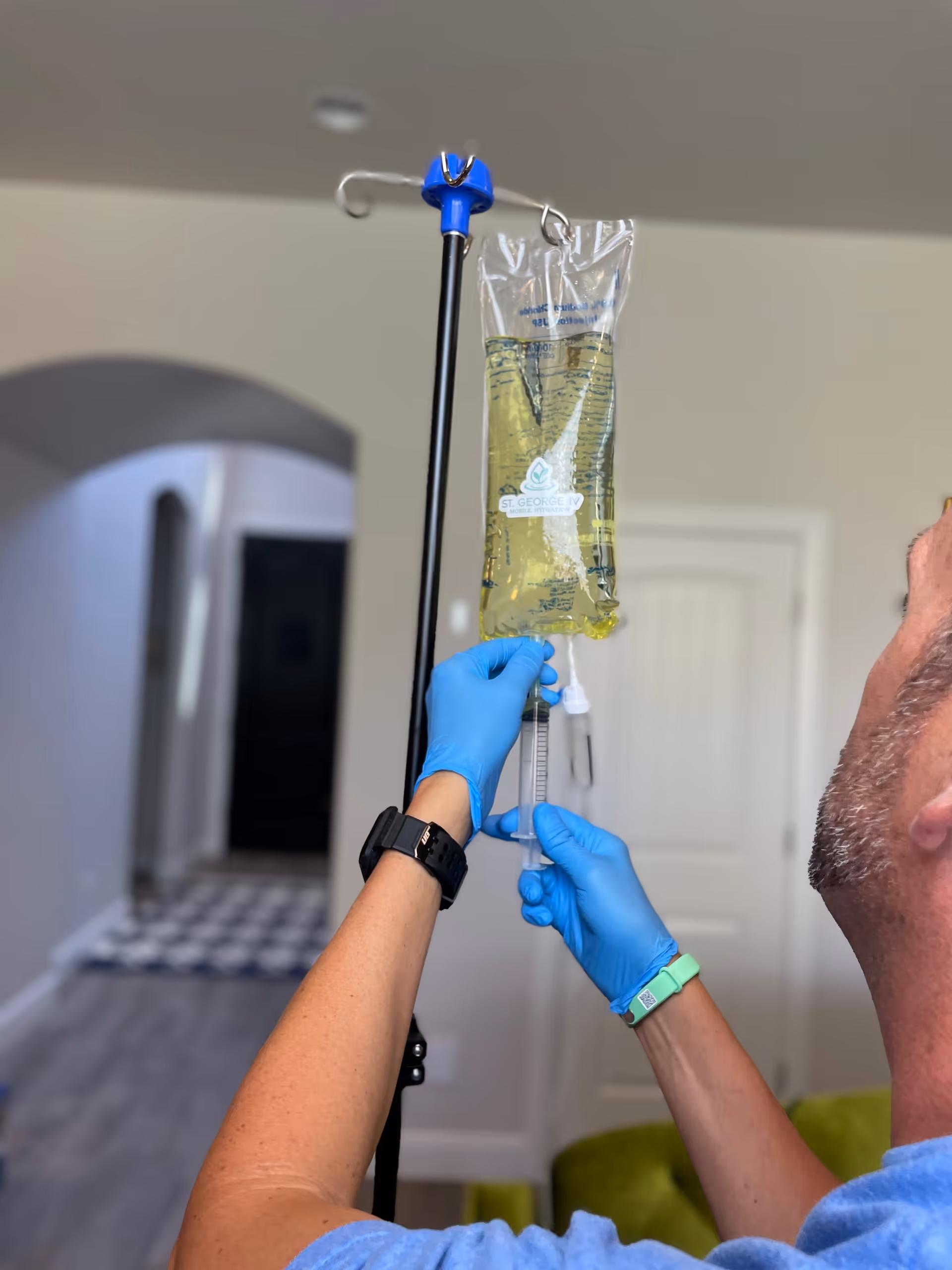 Gloved hands adjust IV bag and syringe on pole inside a senior living facility.