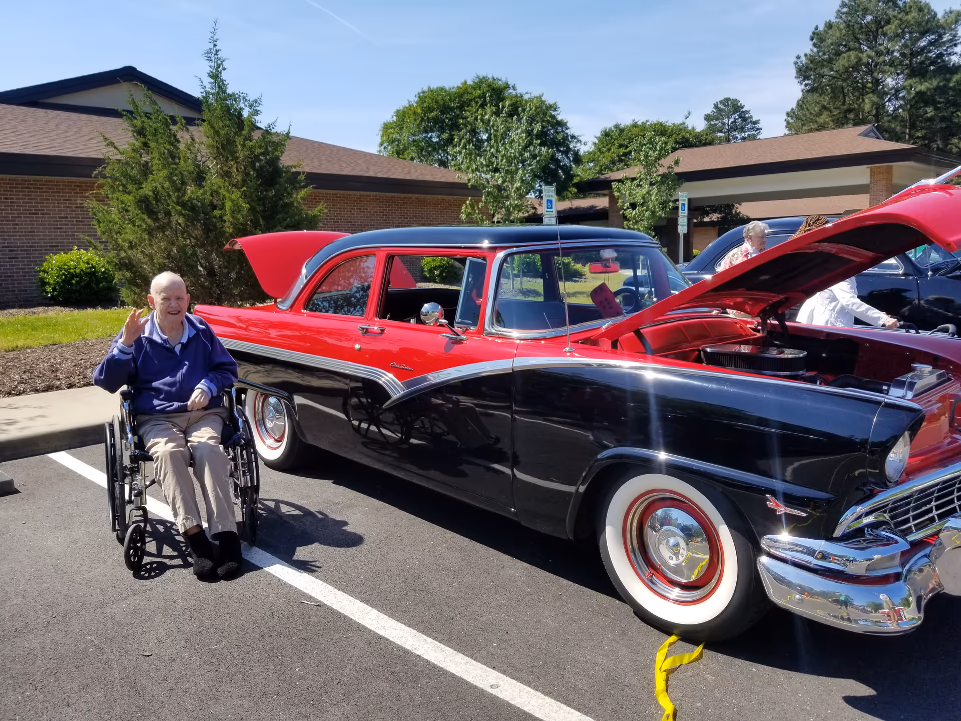 Outdoor scene at nursing facility with an elderly man in a wheelchair beside a vintage car.