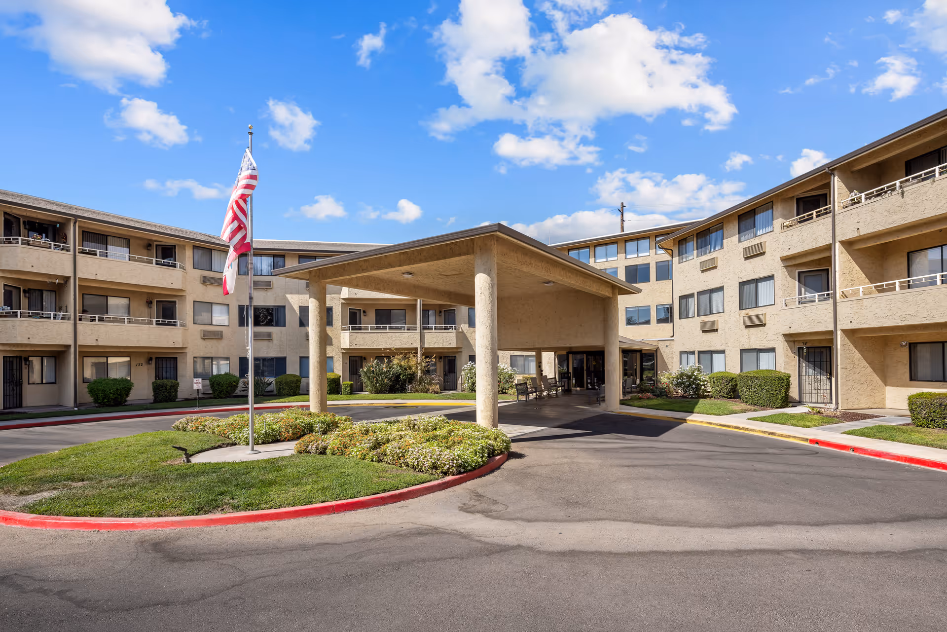 Exterior view of Walnut Park - A Provincial Senior Living Community, showing a three-story beige building with balconies and windows. In front is a covered entrance with columns, a circular driveway, landscaped greenery, and an American flag on a flagpole. The sky is blue with scattered clouds.