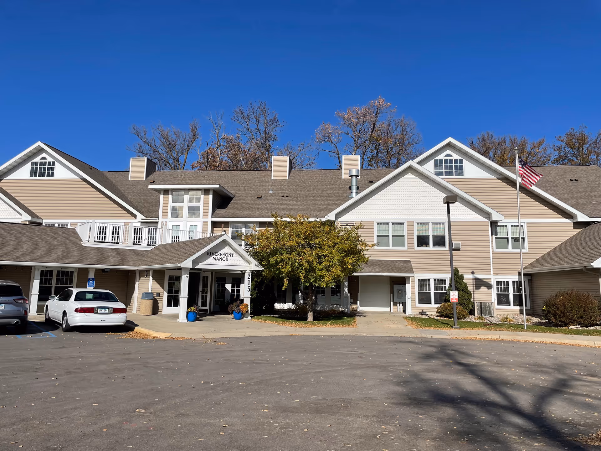 Riverfront Manor senior living building with a front entrance, parked cars, and a clear blue sky.