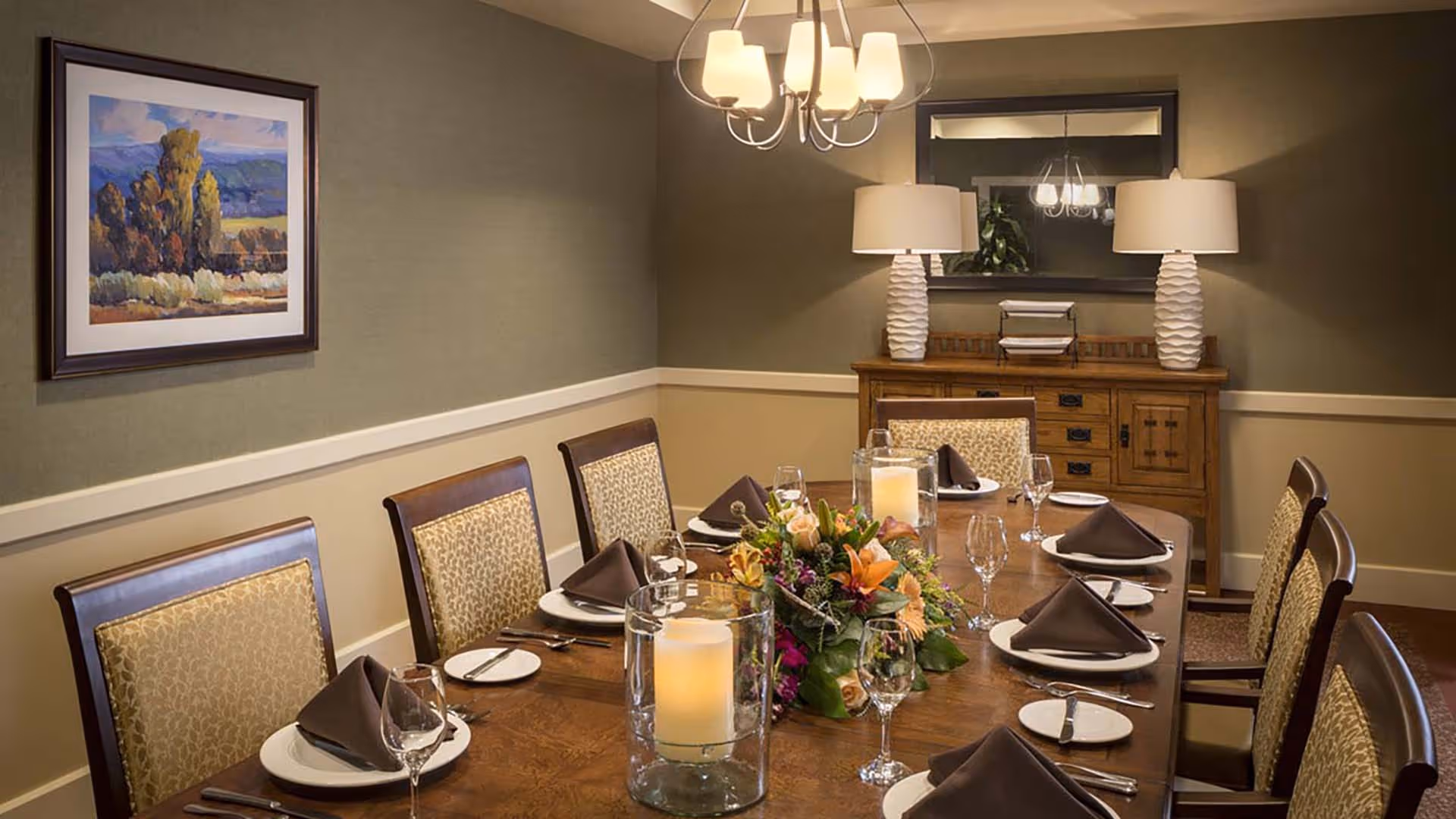 A formal dining room with a wooden table set for eight people. Each place setting includes a white plate, a brown folded napkin, a wine glass, a small bread plate, and silverware. The table is decorated with a floral centerpiece and two large glass candle holders with lit candles. The room has beige and olive green walls, a framed landscape painting on one wall, a wooden sideboard with two white lamps, and a large mirror above it. A chandelier with multiple lights hangs above the table.