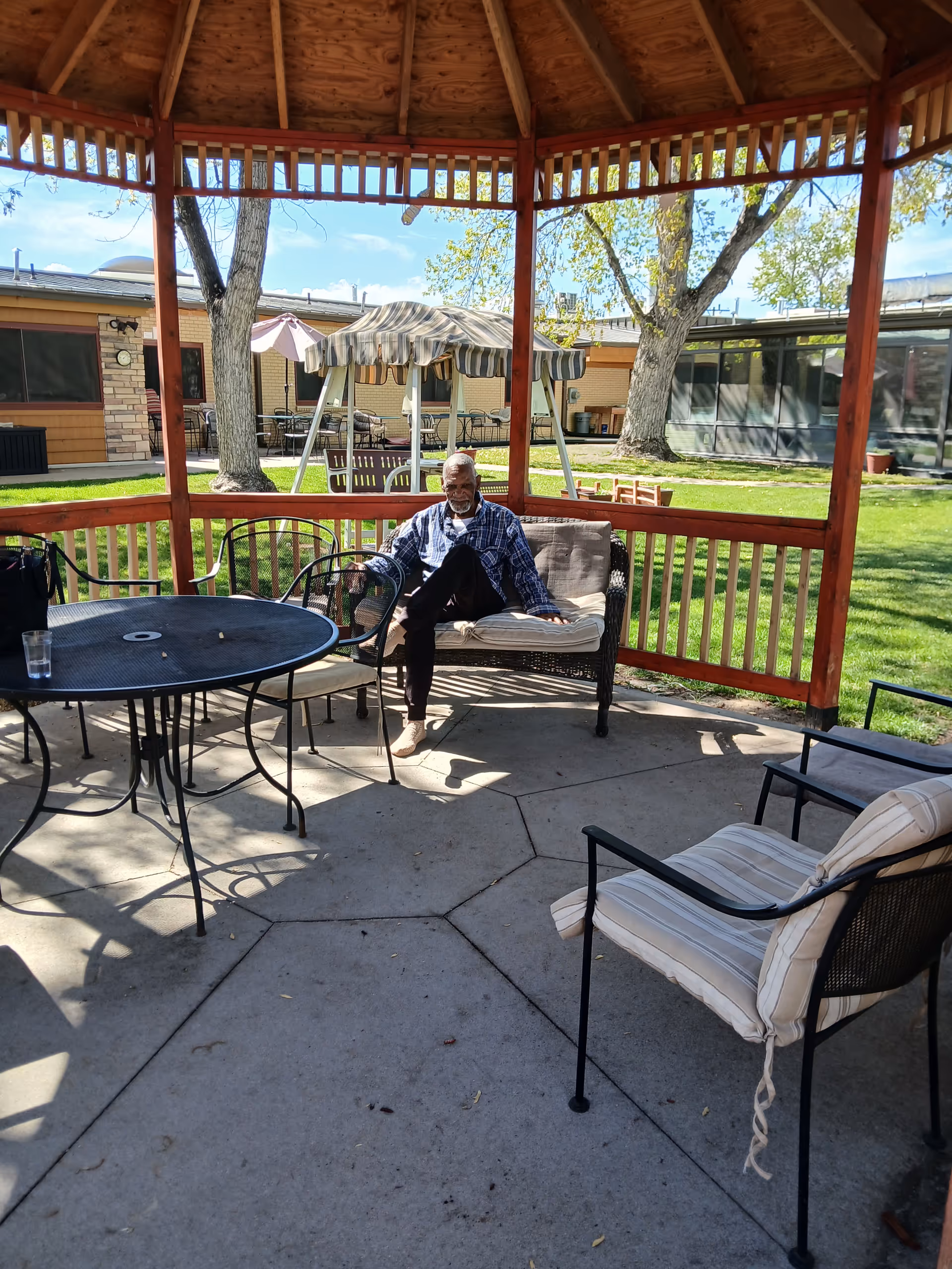 An elderly man sits on a couch under a wooden gazebo in an outdoor courtyard.