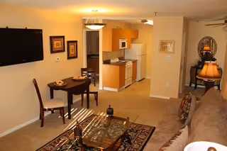 Interior view of a living space in Mountain View Independent Living featuring a beige sofa with patterned cushions, a glass coffee table on a decorative rug, a small dining table with two chairs, a wall-mounted flat screen TV, and a kitchen area with wooden cabinets, a microwave, refrigerator, and stove in the background.