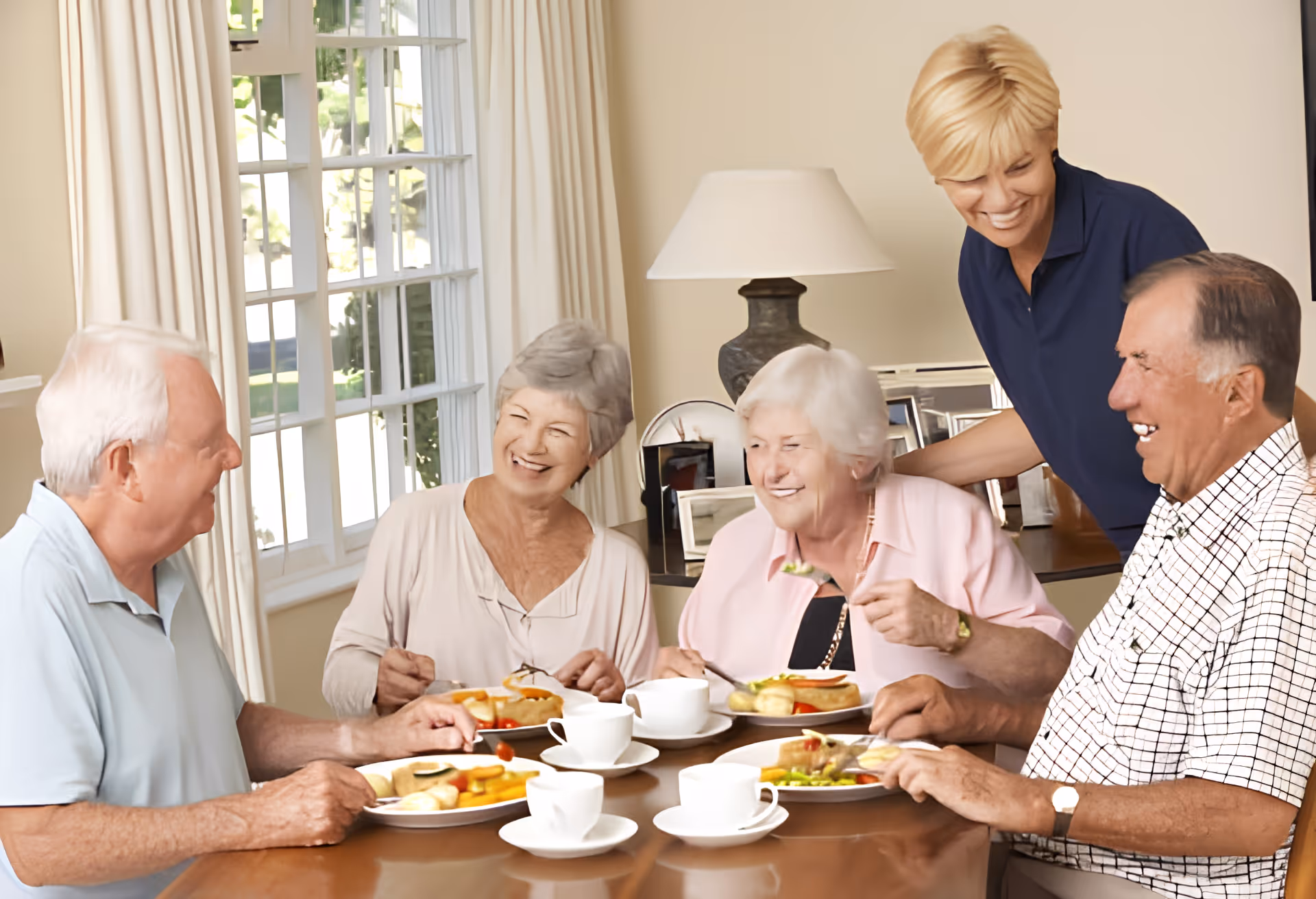 Four elderly people sitting around a dining table enjoying a meal together, with a caregiver standing behind them smiling. The room has a large window with white curtains and a table lamp in the background.