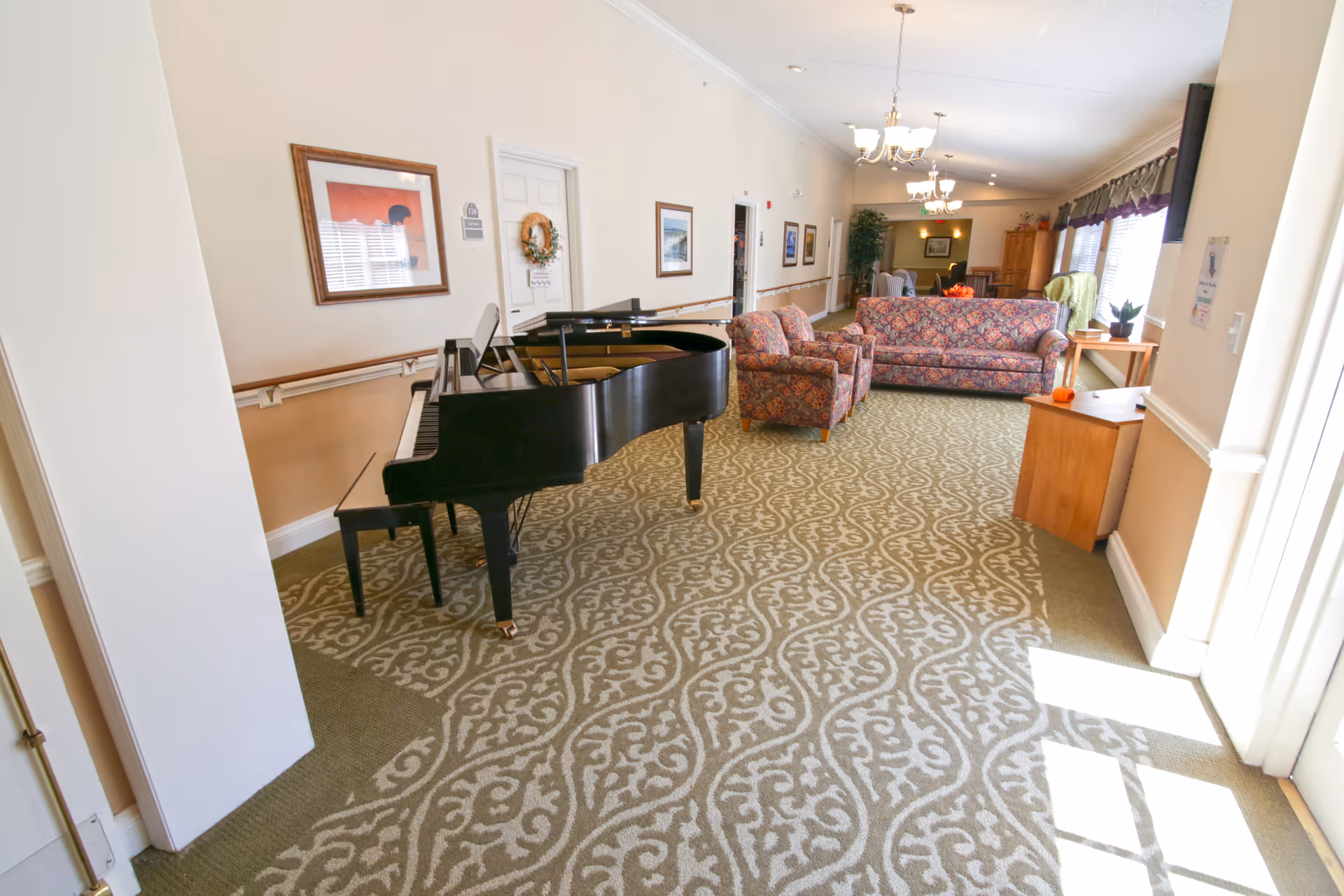 Bright senior living lounge with a grand piano, patterned carpet, and seating area.