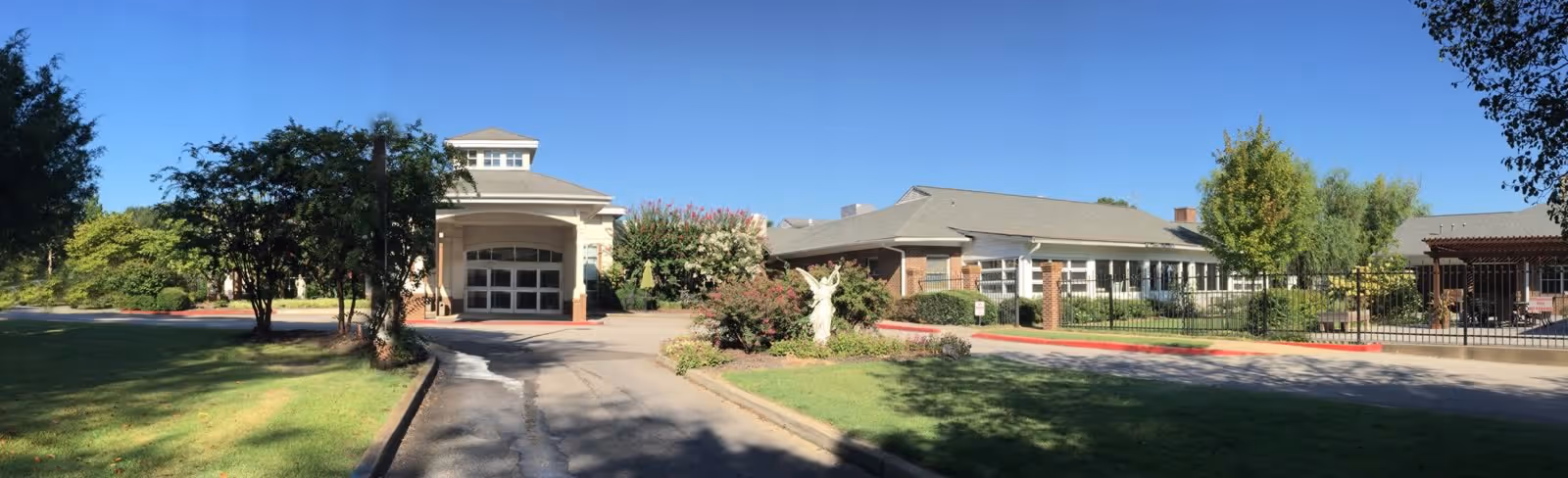 Front exterior view of Ave Maria Home Assisted Living facility with a driveway, green lawn, trees, bushes, and a statue in the center garden area under a clear blue sky.