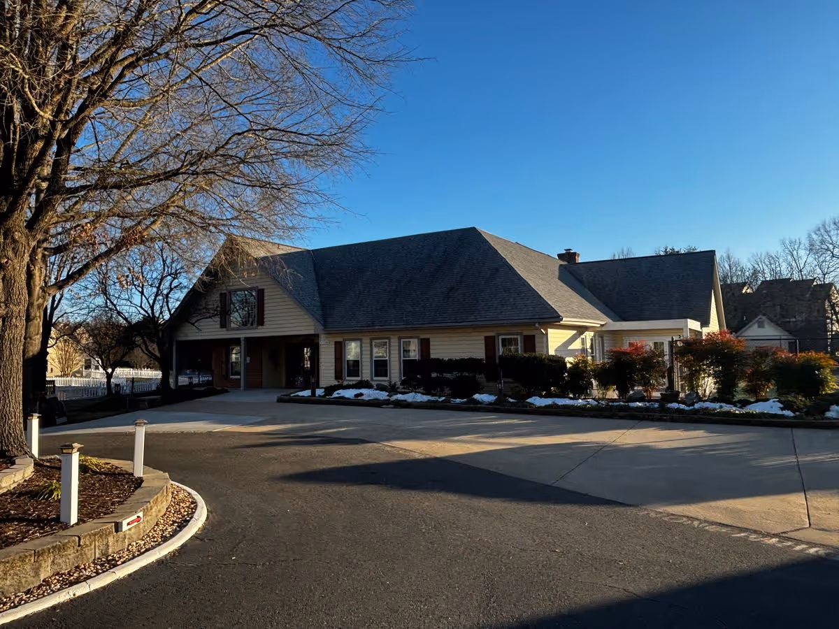Exterior front view of a single-story residential-style building with a pitched roof, driveway, trees, and landscaped beds with patches of snow.
