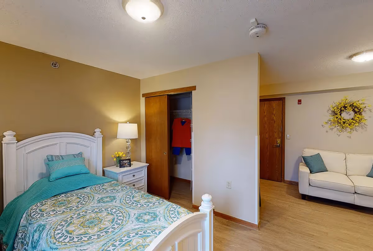 Furnished assisted living bedroom with a white bed and patterned teal bedding, a nightstand and lamp, an open closet, and a small sitting area with a white sofa.