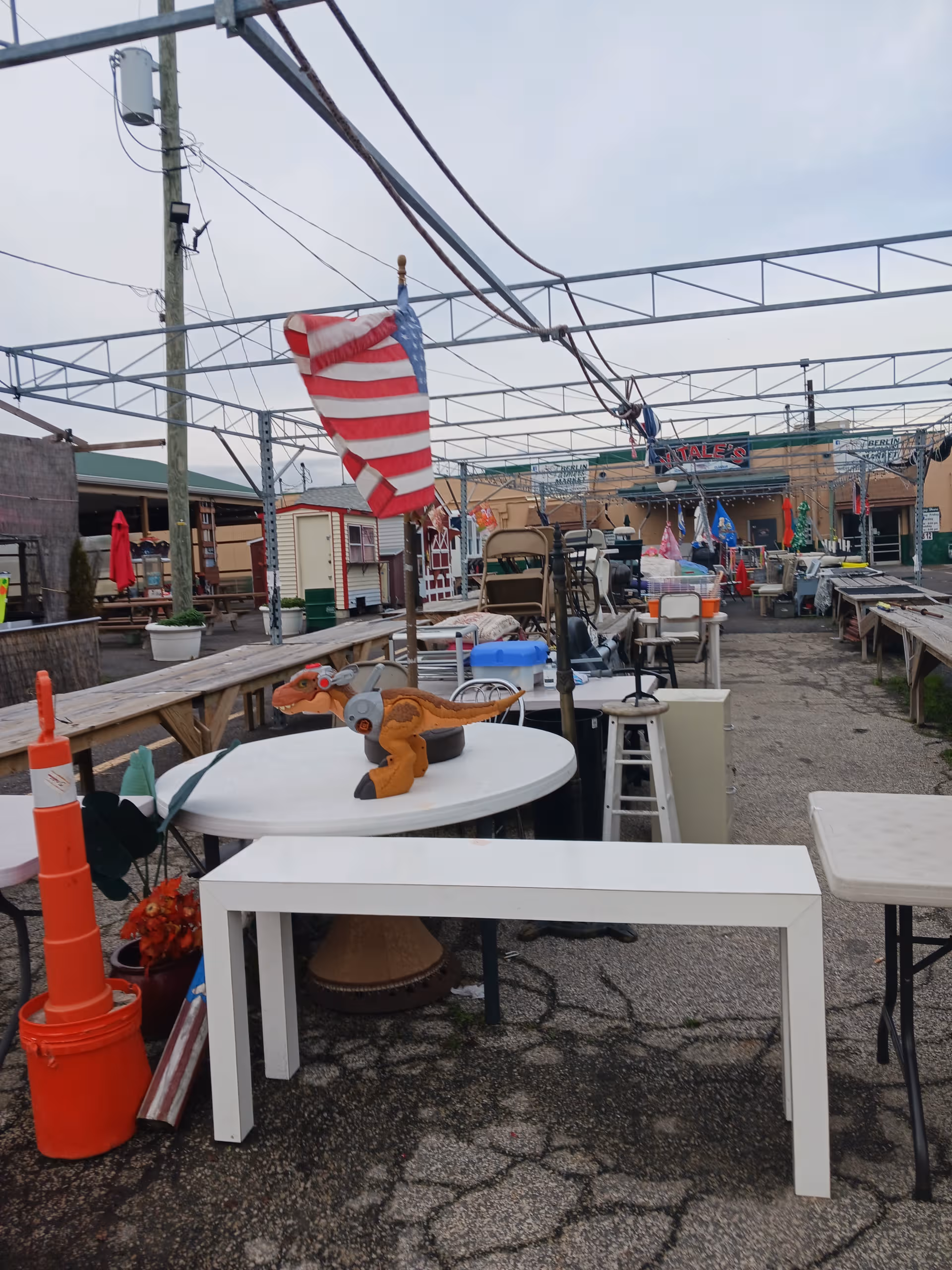 Outdoor flea market scene with tables, chairs, items, and an American flag under a cloudy sky.