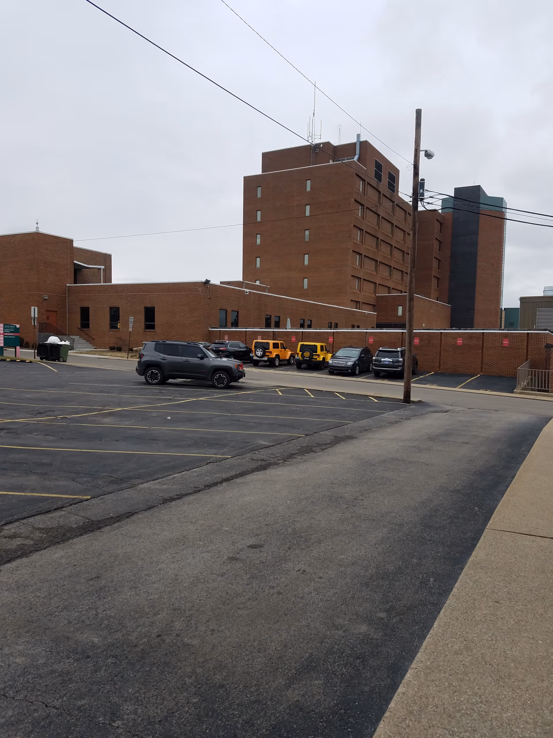 Brick hospital-like building with a parking lot and few cars under an overcast sky.