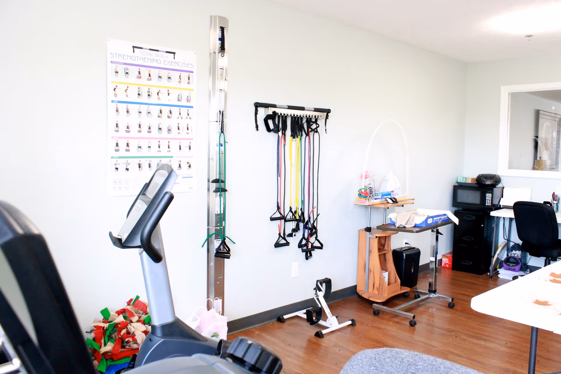 Small fitness/therapy room with exercise equipment, resistance bands hanging on the wall, a treadmill, and a desk area.