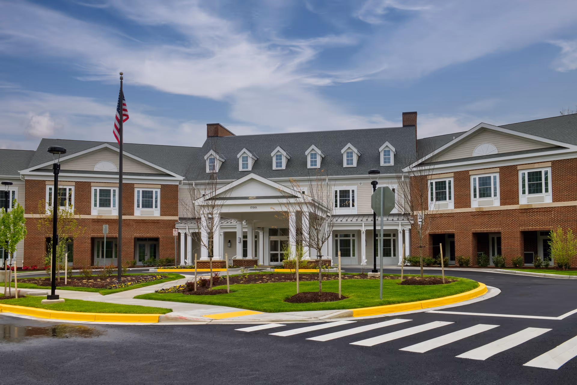 Front exterior view of a large senior living facility building with brick and white siding, multiple windows, a covered entrance, landscaped greenery, a flagpole with an American flag, and a crosswalk in the foreground under a partly cloudy sky.