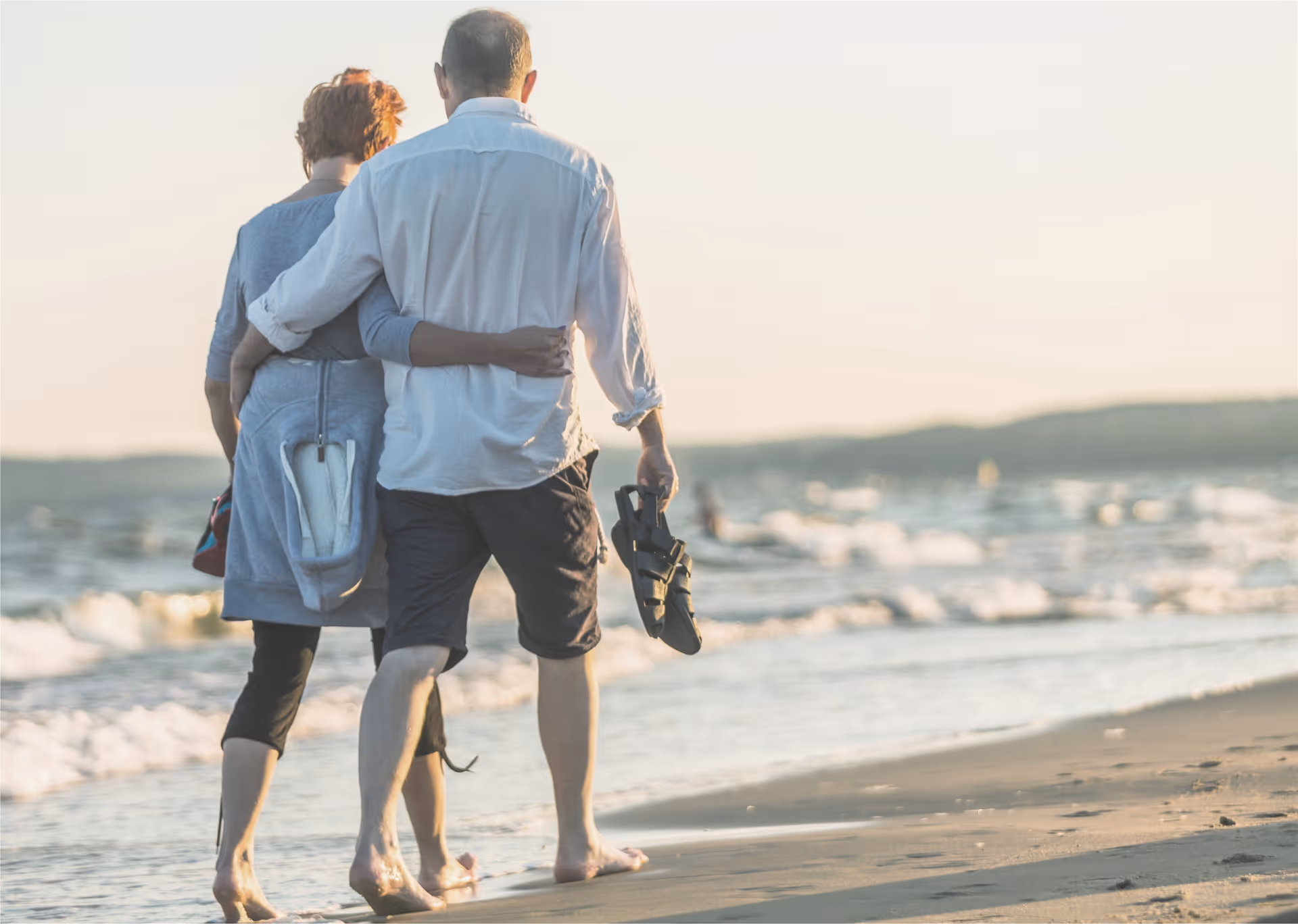Couple strolls along the beach, arm-in-arm, barefoot, sandals in hand at sunset.