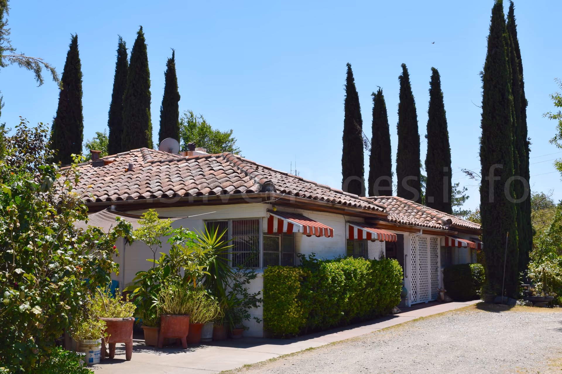 Single-story retirement home exterior with terracotta tile roof, striped awnings, and tall cypress trees.