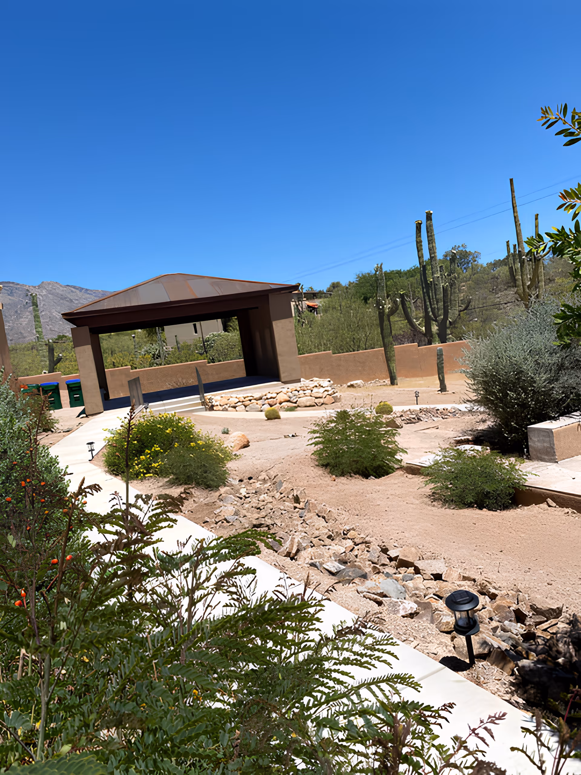Desert courtyard with a covered pavilion, walking paths, cacti and shrubs under a clear blue sky.