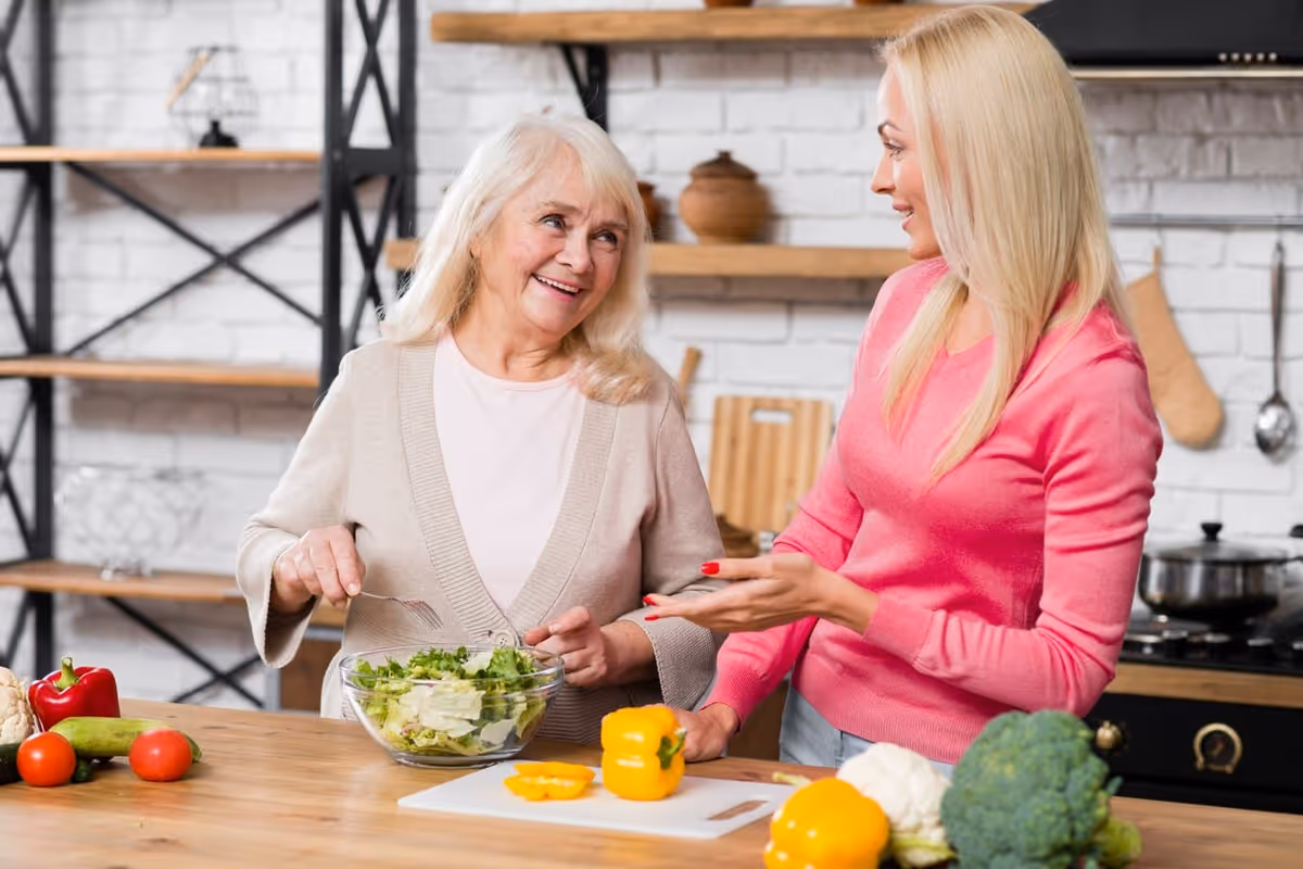 An older woman and a younger woman smiling as they prepare a salad together in a modern kitchen.