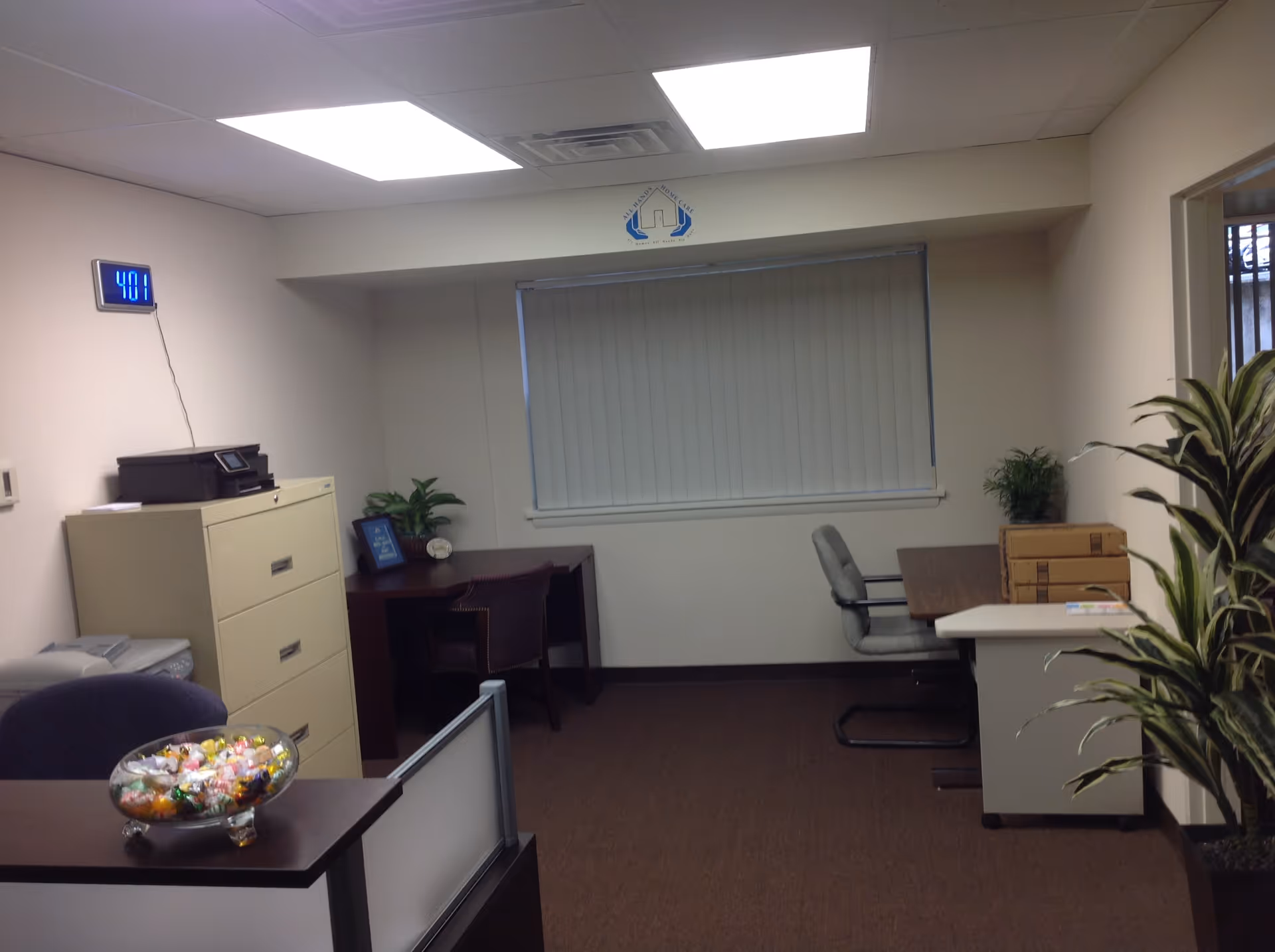 An indoor office area with desks, a window, filing cabinets, printers, and potted plants.