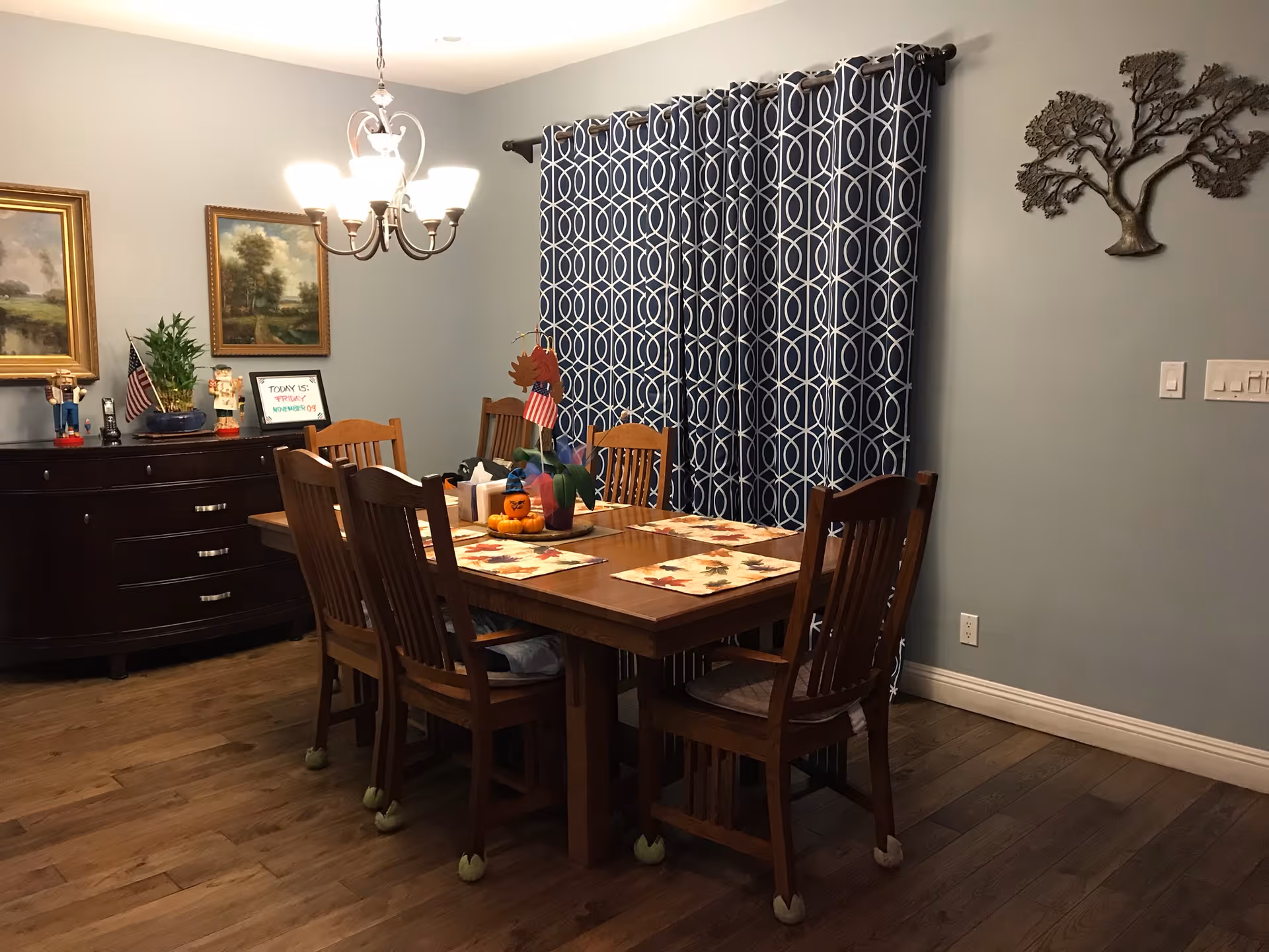 A dining room with a wooden table set for six with fall-themed placemats and a centerpiece featuring a small pumpkin and an American flag. The room has wooden chairs with tennis balls on the legs, a dark wooden sideboard with decorative items and framed paintings on the wall, a chandelier overhead, and blue curtains with a white geometric pattern covering a window.