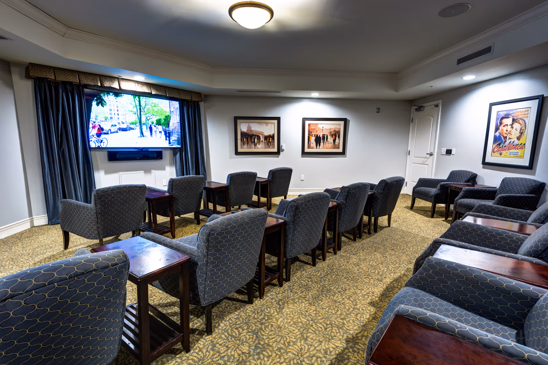 Carpeted common room with rows of upholstered chairs and side tables facing a wall-mounted TV and framed artwork.