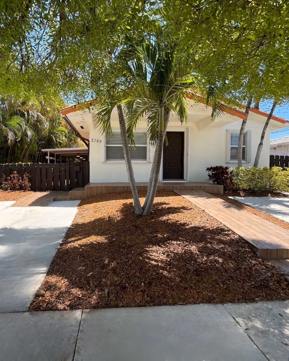 Front exterior view of a single-story white house with a brown door, two windows with blinds, and a small porch. There is a palm tree in the mulched front yard, a concrete driveway on the left, and a wooden ramp leading to the porch. The house number 2789 is visible on the wall near the door. Surrounding the house are green trees and bushes under a clear blue sky.