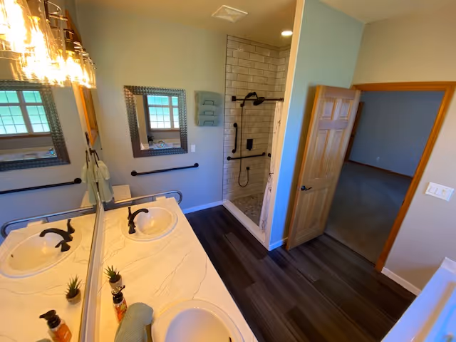 Bright shared bathroom with double sinks, mirrors, grab bars, and a glass-enclosed walk-in shower.