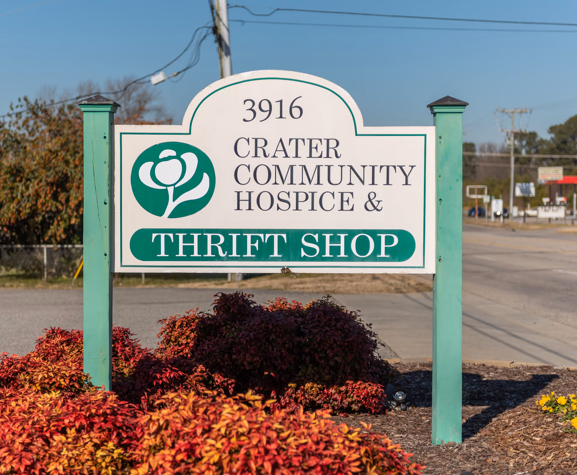 A white sign reading Crater Community Hospice & Thrift Shop, with green posts and red shrubs.