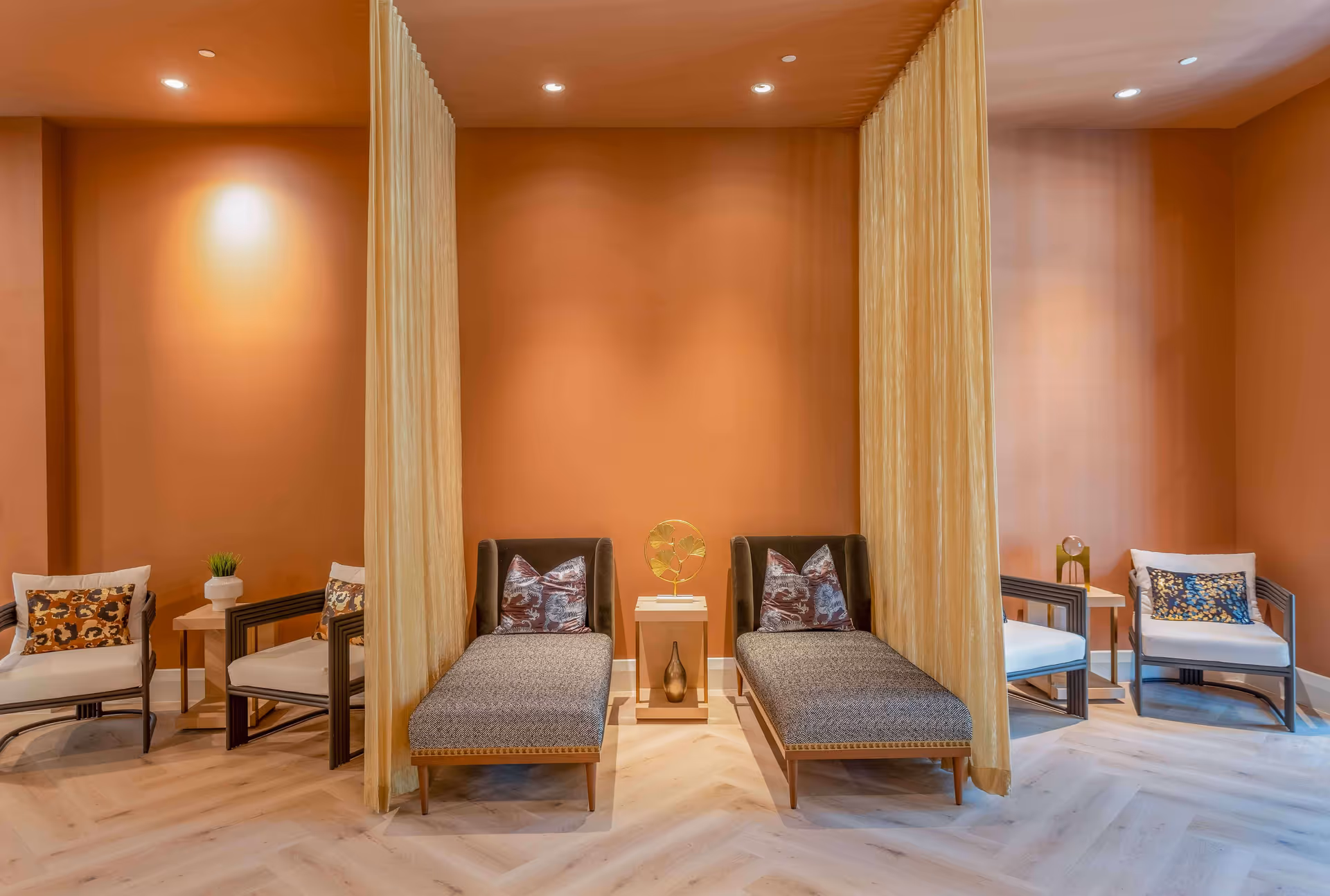 Warm-toned communal lounge with two upholstered chaise lounges separated by sheer curtains and armchairs on either side.