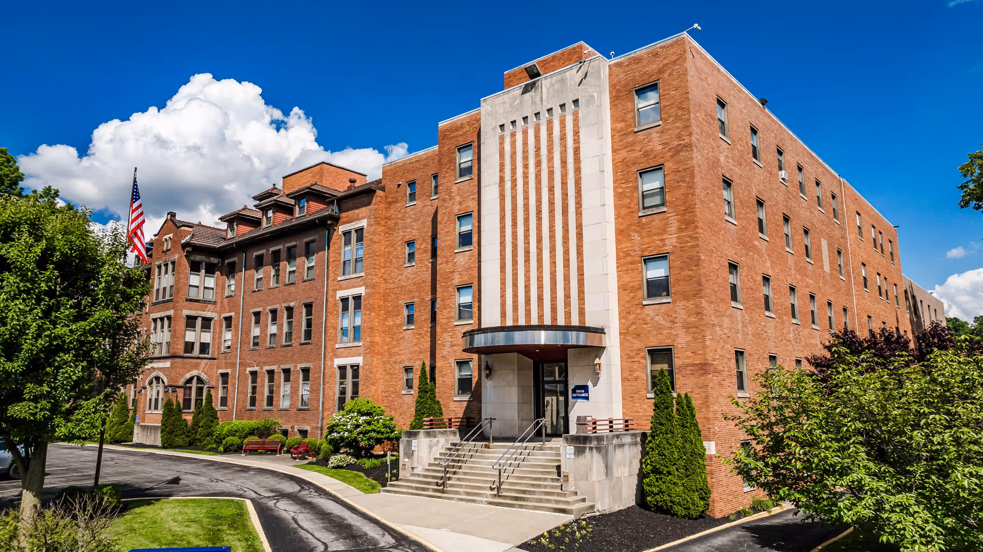 Brick multi-story senior care building front with a main entrance, steps, American flag and landscaped grounds under a blue sky.