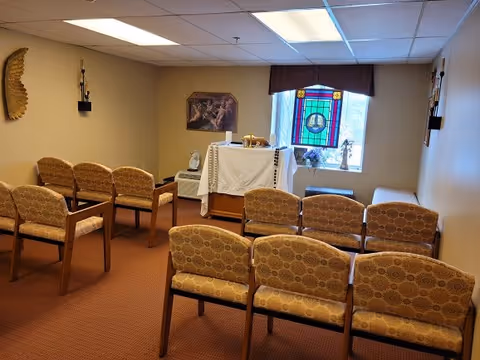 Small chapel-like interior with rows of patterned upholstered chairs facing a covered altar beneath a stained-glass window.