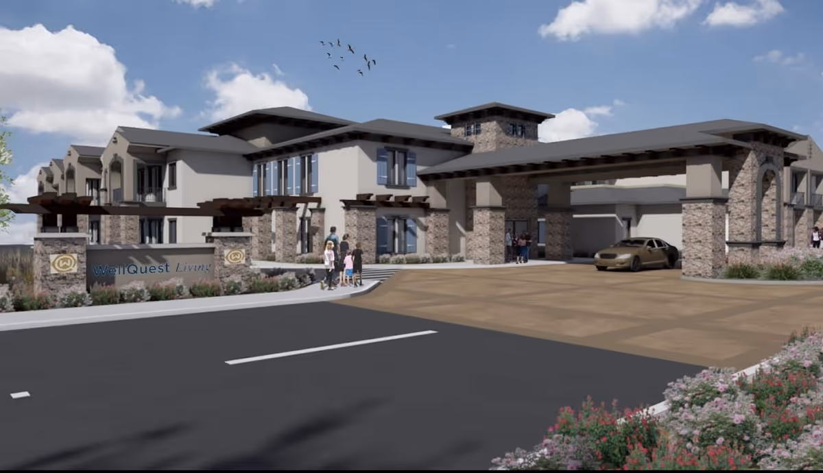 Exterior view of the WellQuest of Granite Bay senior living facility entrance with a porte-cochere, stone columns, landscaping, a sign reading 'WellQuest Living', and people near the driveway.