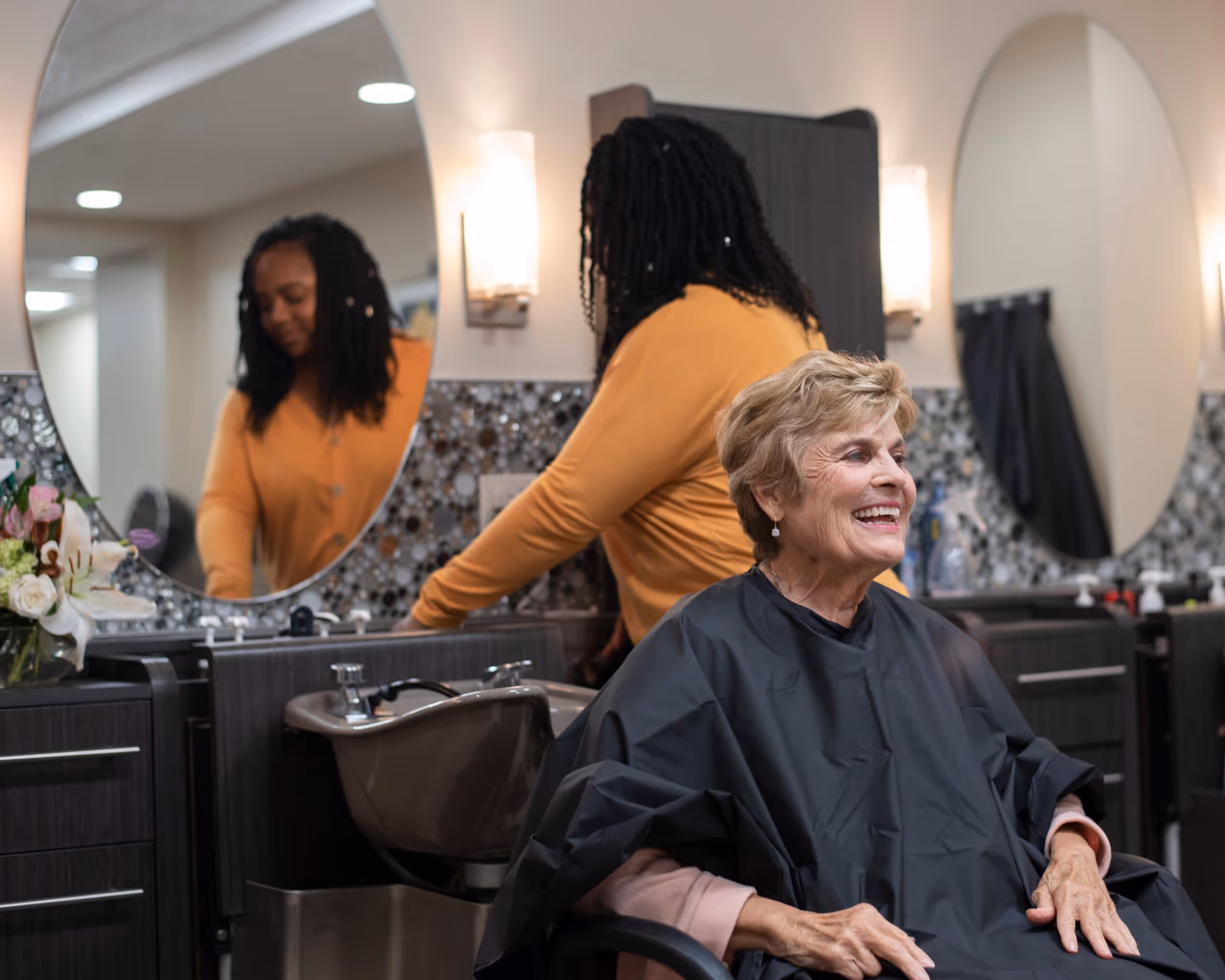 An elderly woman with short blonde hair sits in a salon chair wearing a black cape, smiling. Behind her, a hairstylist with long braided hair and an orange sweater is preparing to wash hair at a salon sink. The background features two large oval mirrors and mosaic tile backsplash with wall sconces providing light.