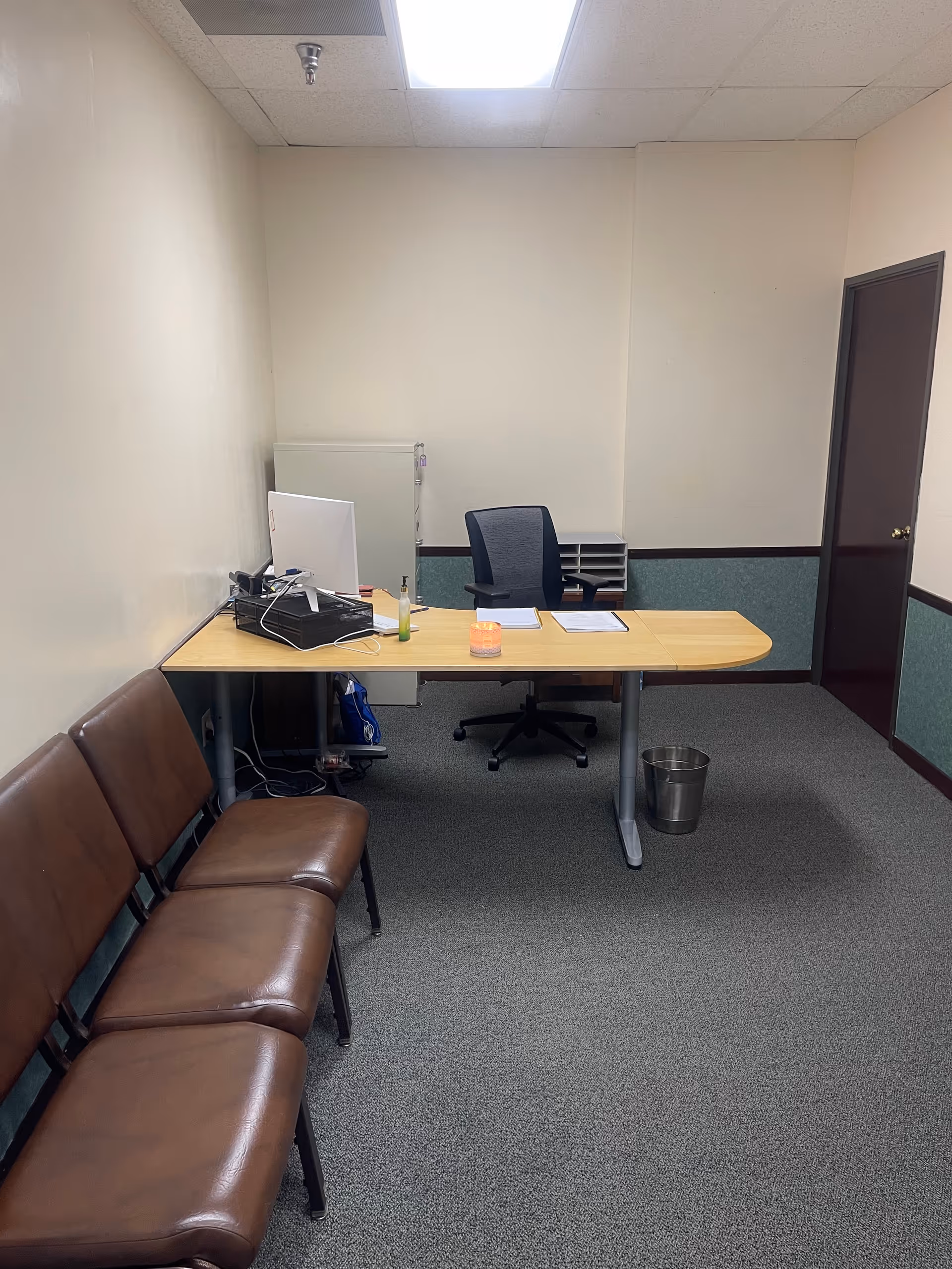 Office desk with computer and papers, white walls, brown chairs along the left wall.