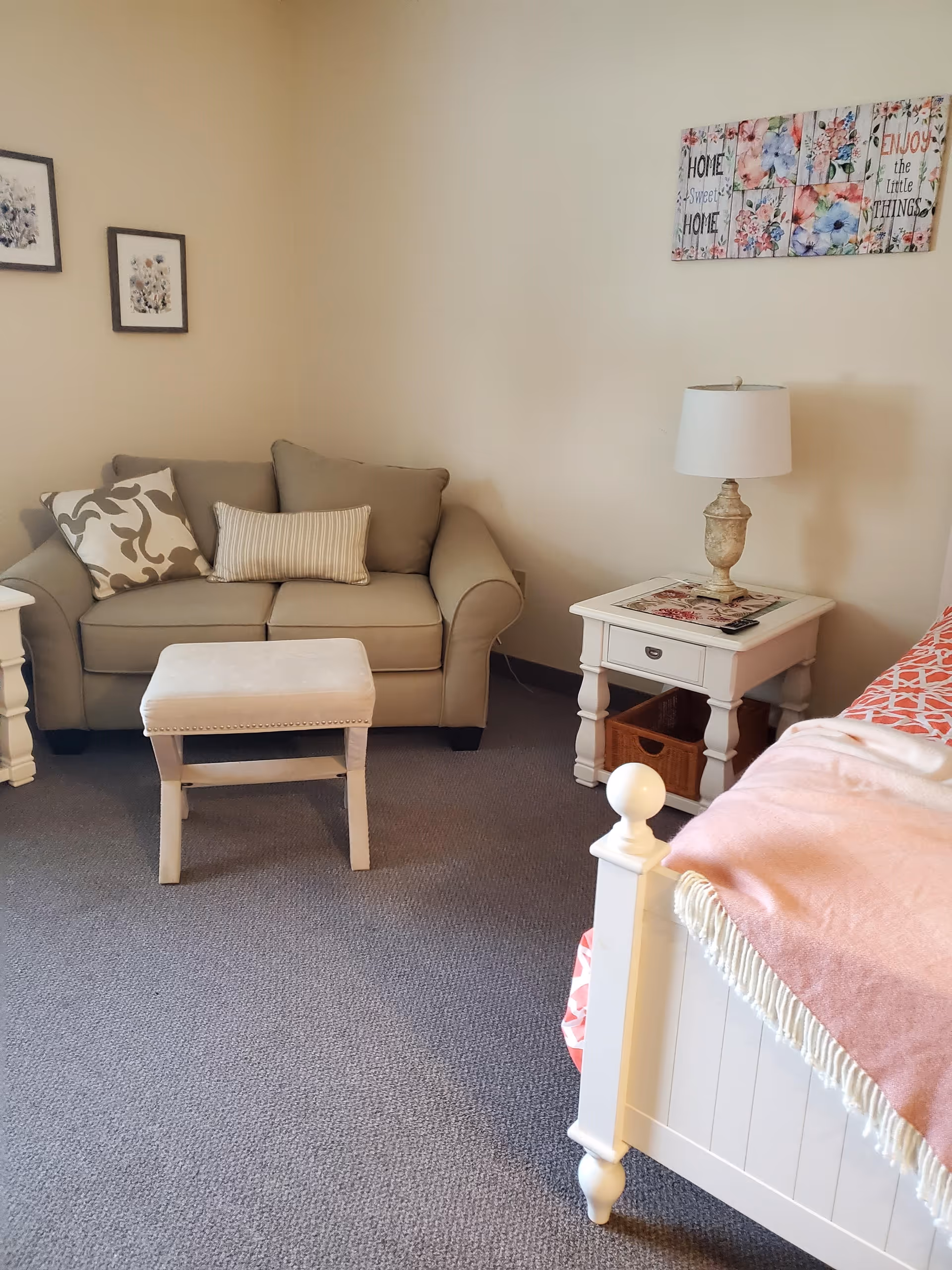 A cozy room with a beige loveseat adorned with two decorative pillows, a small cushioned ottoman in front of it, a white wooden nightstand with a lamp and a basket underneath, and a white bed with a pink blanket partially visible. The walls are decorated with framed floral artwork and a colorful sign that reads 'Home Sweet Home' and 'Enjoy the Little Things.'
