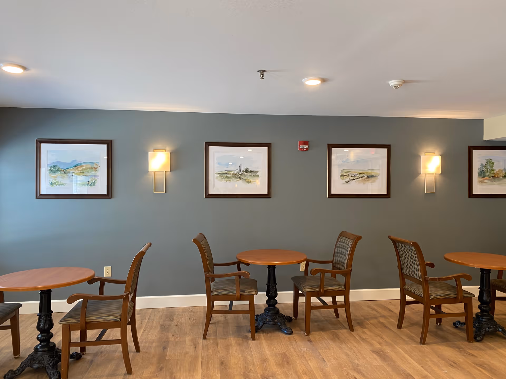 A seating area with three round wooden tables, each accompanied by two wooden chairs with armrests and patterned cushions. The room has a light wooden floor and a muted green wall adorned with four framed landscape paintings and two wall-mounted light fixtures emitting a warm glow.