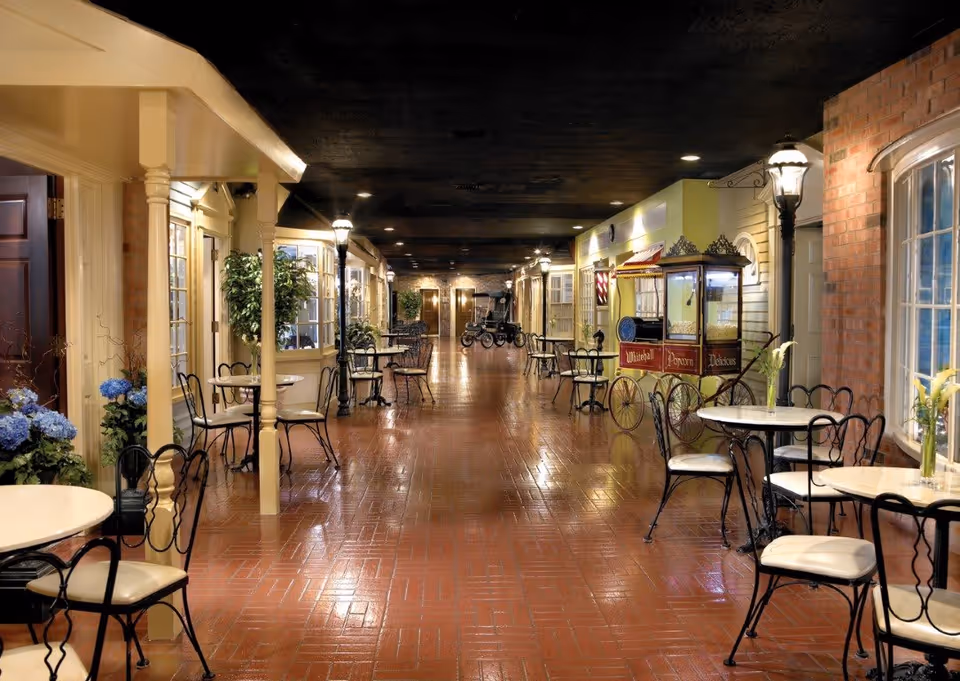 Indoor hallway with a polished brick floor, lined with small round tables and black wrought iron chairs with white cushions. The hallway is decorated with potted plants, streetlamp-style lights, and a vintage popcorn cart labeled 'Whitehall Popcorn Delicious'. Doors and windows resembling storefronts or rooms line both sides of the hallway.